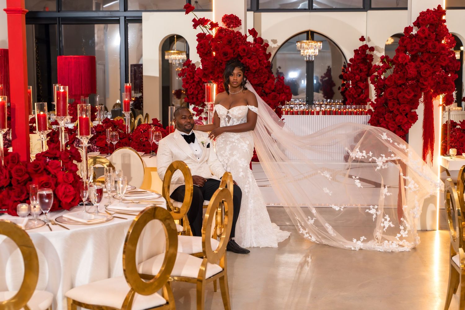 Wedding reception venue decorated with dramatic red floral arrangements and gold chairs at white-clothed tables.