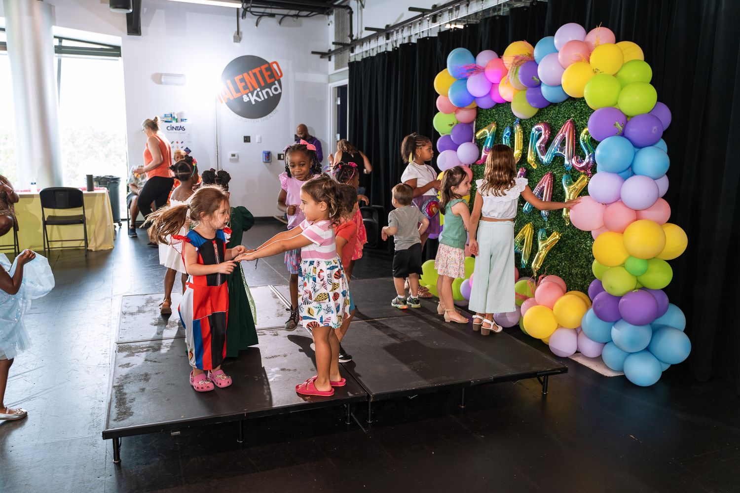 Kids in costumes performing on stage with colorful balloon decorations.