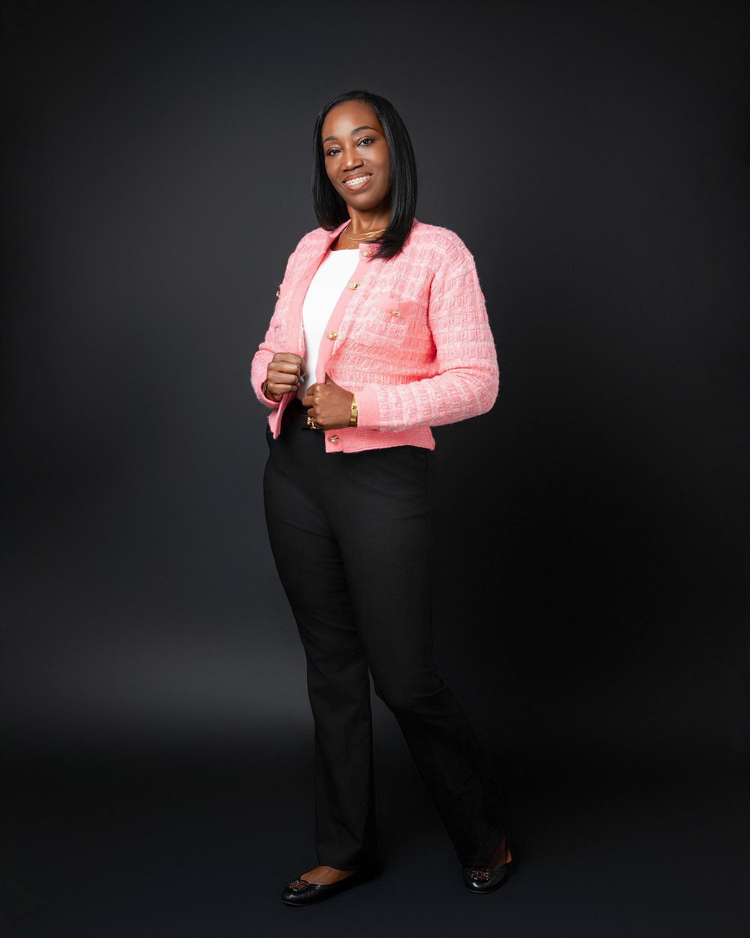 Professional portrait of someone wearing a coral pink blazer and black pants against a dark background.