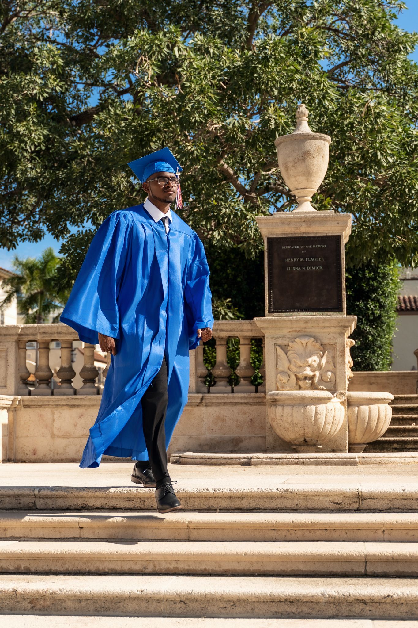 A graduate in bright blue cap and gown walks down stone steps next to an ornate decorative urn on a sunny day.