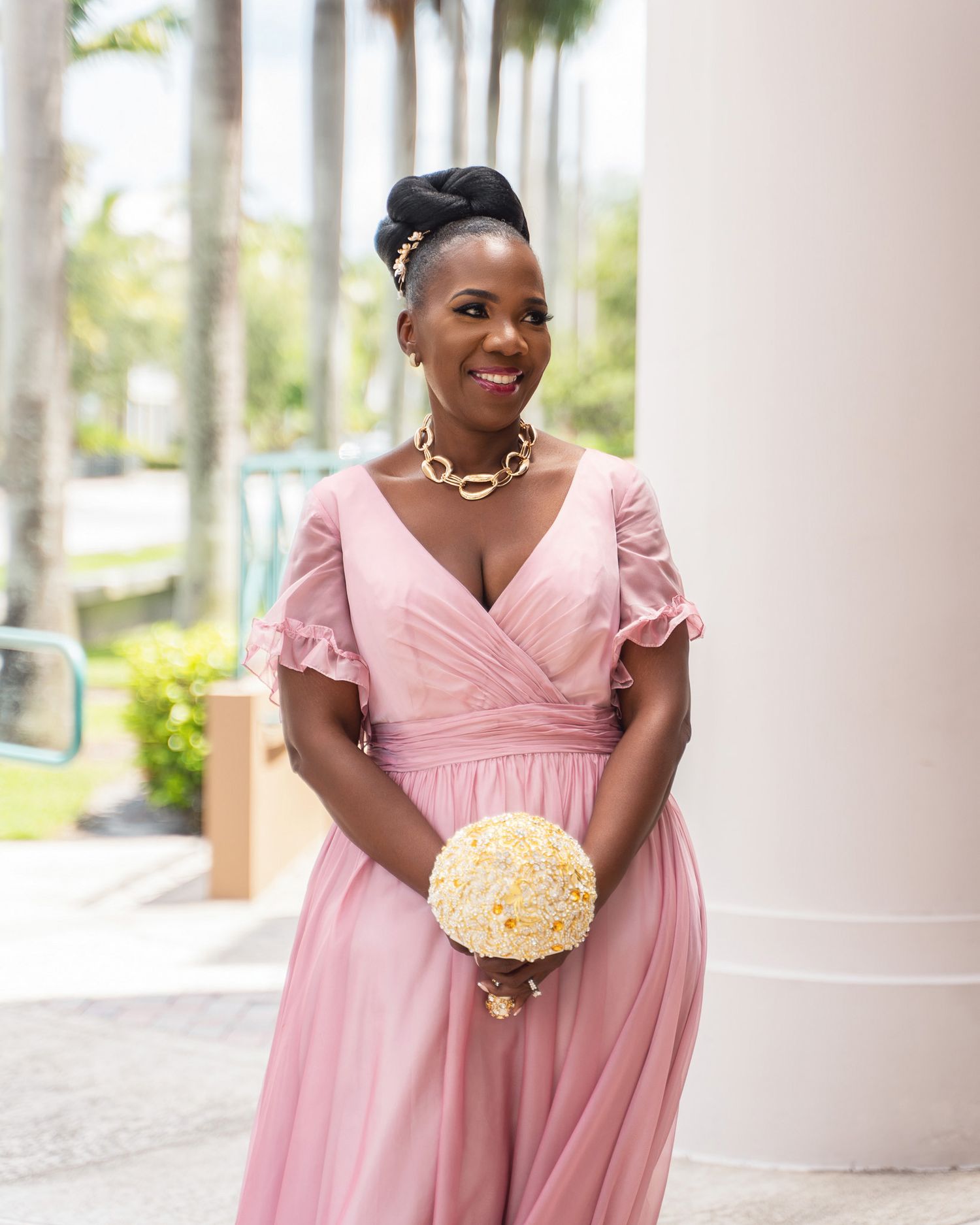 Woman in pink dress holds cream-colored bouquet against tropical backdrop.