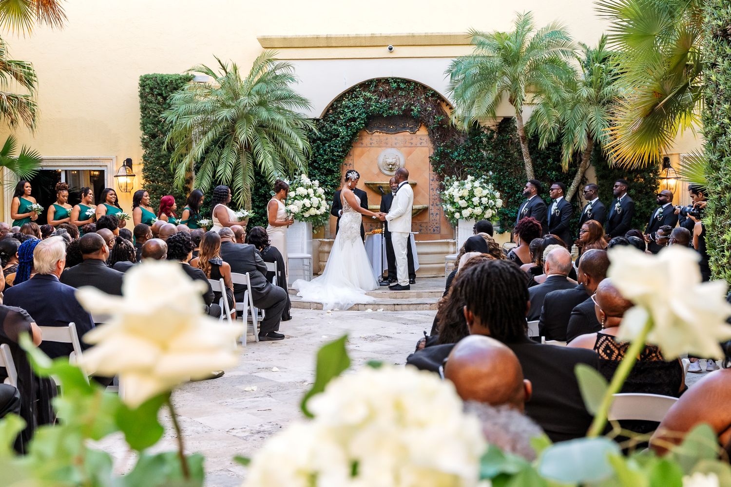 Outdoor wedding ceremony with palm trees and white floral decorations in a courtyard setting.
