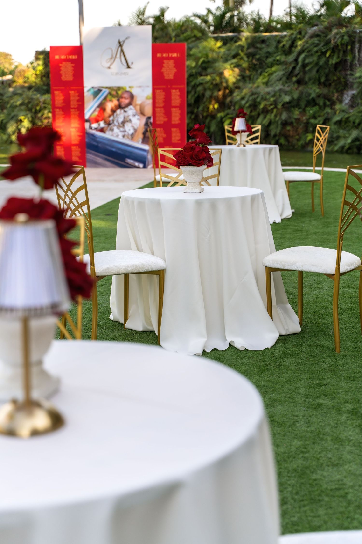 Outdoor reception area with white round tables and gold chairs set up on grass with red accents.