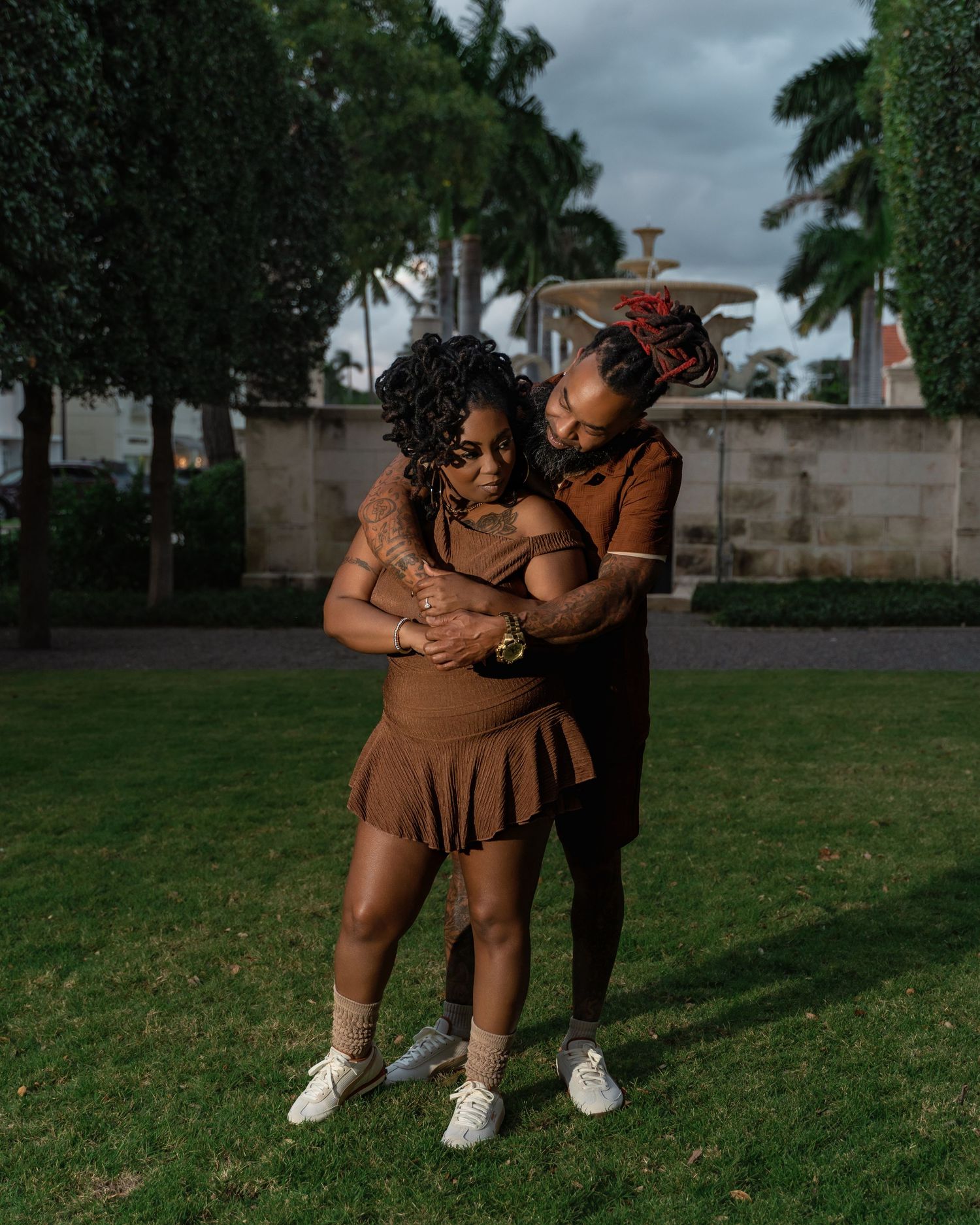 A couple in brown outfits dances together on a grassy lawn with palm trees and buildings in the background.
