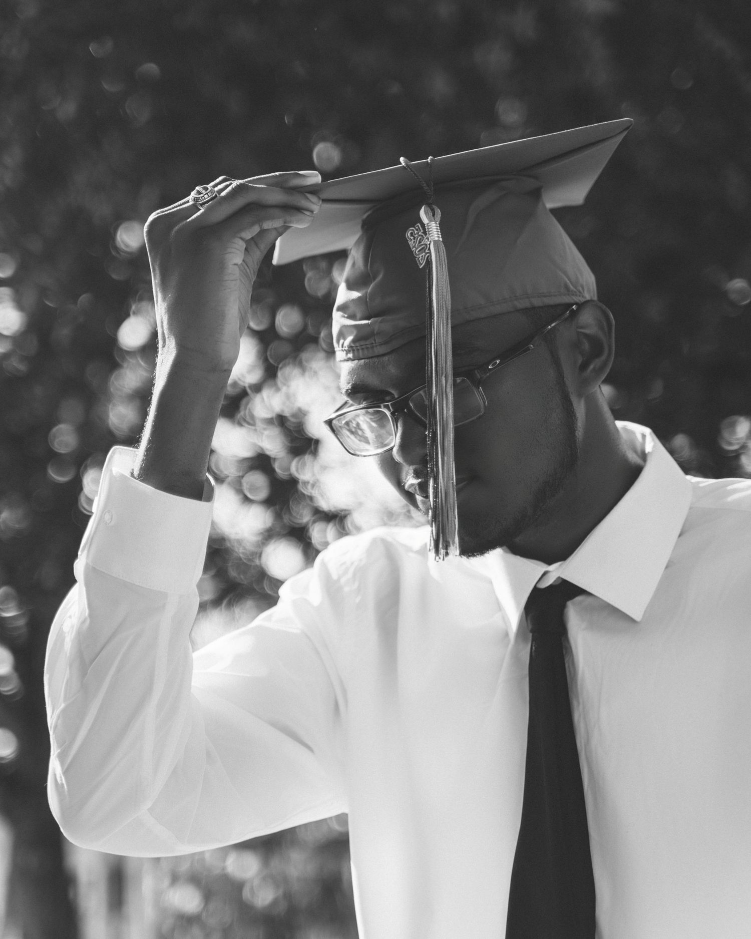 A graduate in white dress shirt and tie adjusts their mortarboard cap in a black and white portrait with bokeh background.
