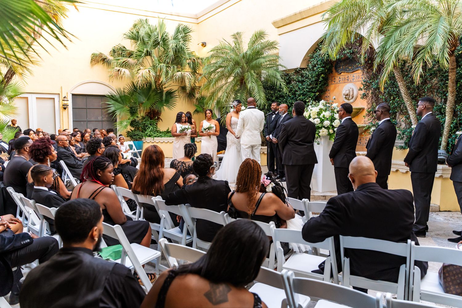 Wedding ceremony setup with white chairs and tropical landscaping in an elegant courtyard.