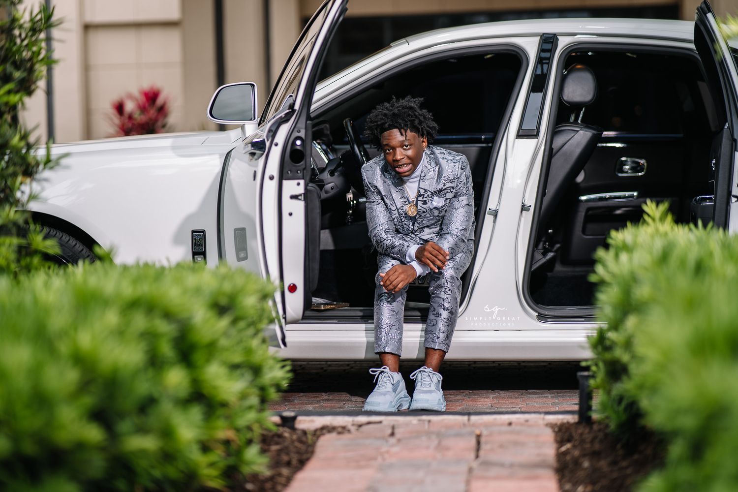 A person in a silver suit sits in the doorway of a white luxury car.