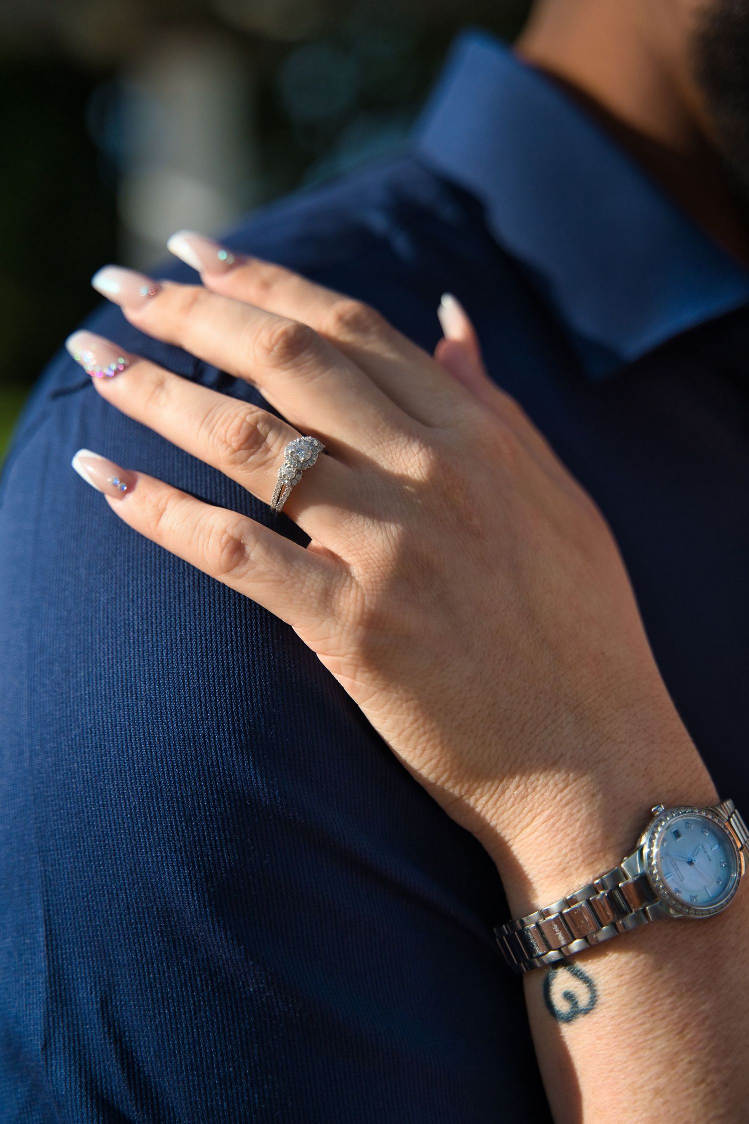 Close-up of hands showing an engagement ring and watch against navy blue fabric.