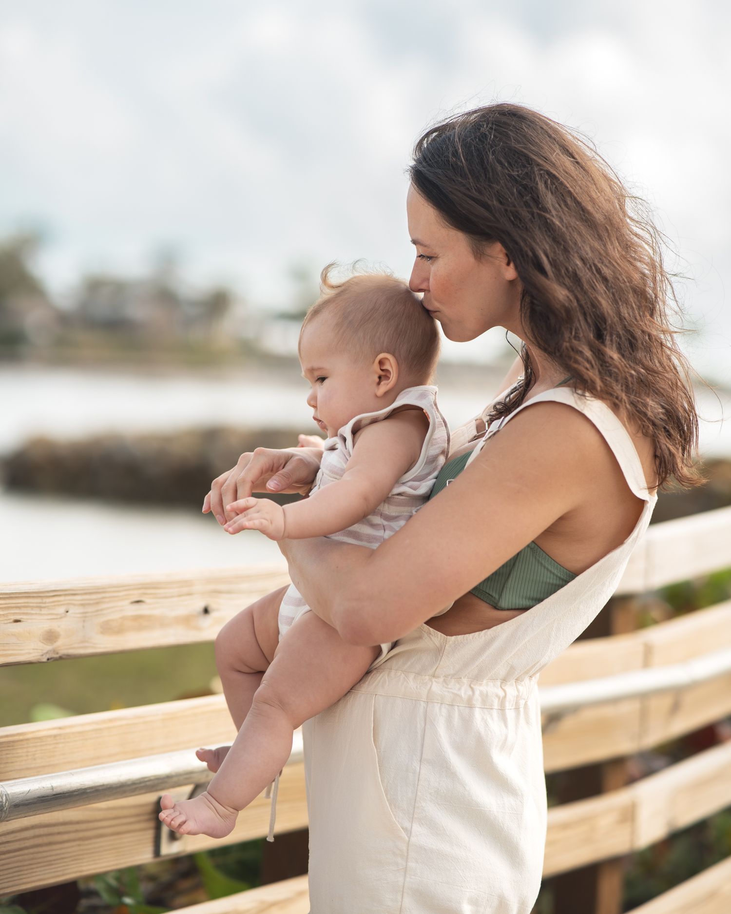 A parent holds a baby while standing at a wooden railing overlooking a coastal view.