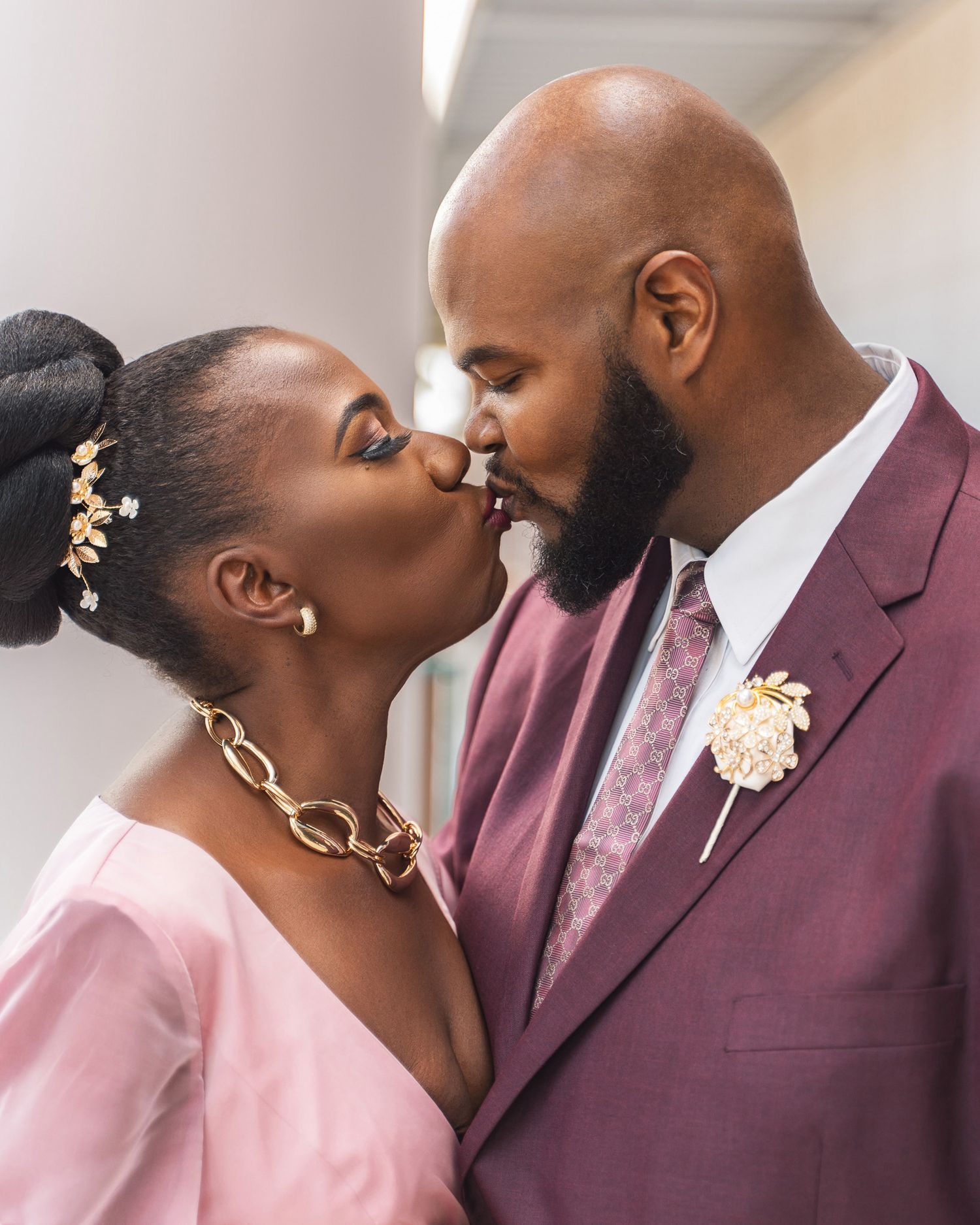 A romantic couple shares a tender kiss while wearing coordinating pink and burgundy formal attire.