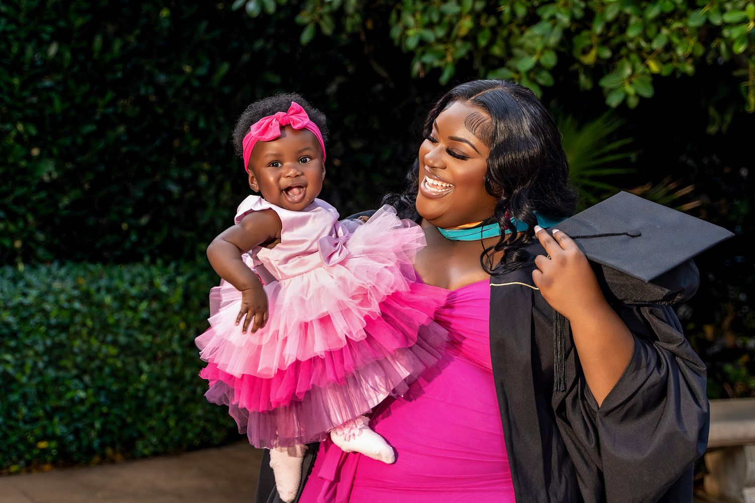 Graduate holds a baby wearing a pink tulle dress against a garden backdrop during celebration.