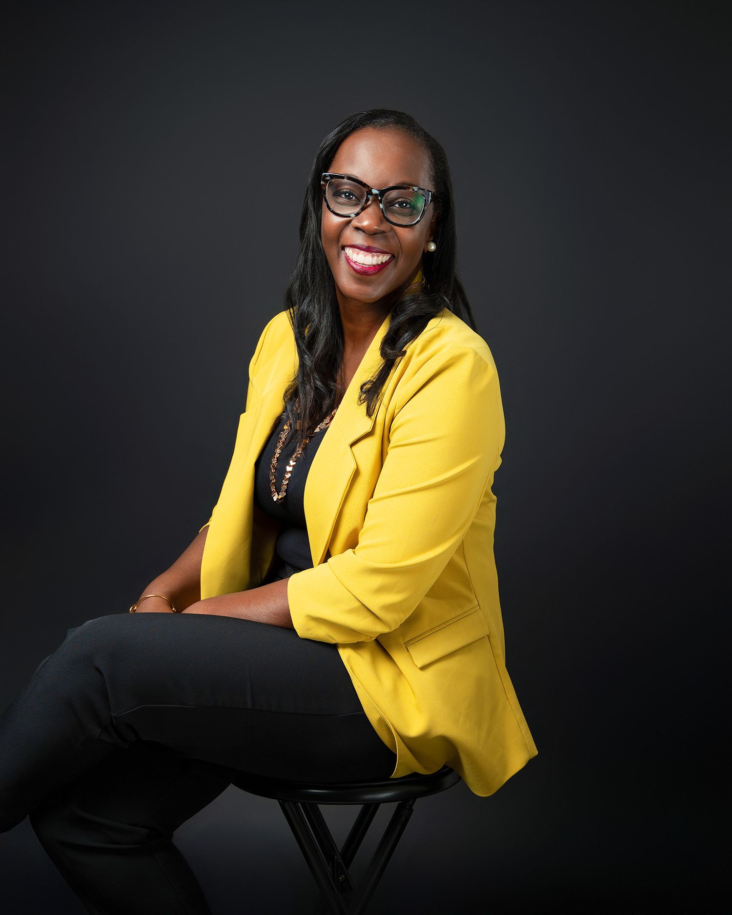 Studio portrait of someone in a bright yellow blazer seated on a stool against dark background.