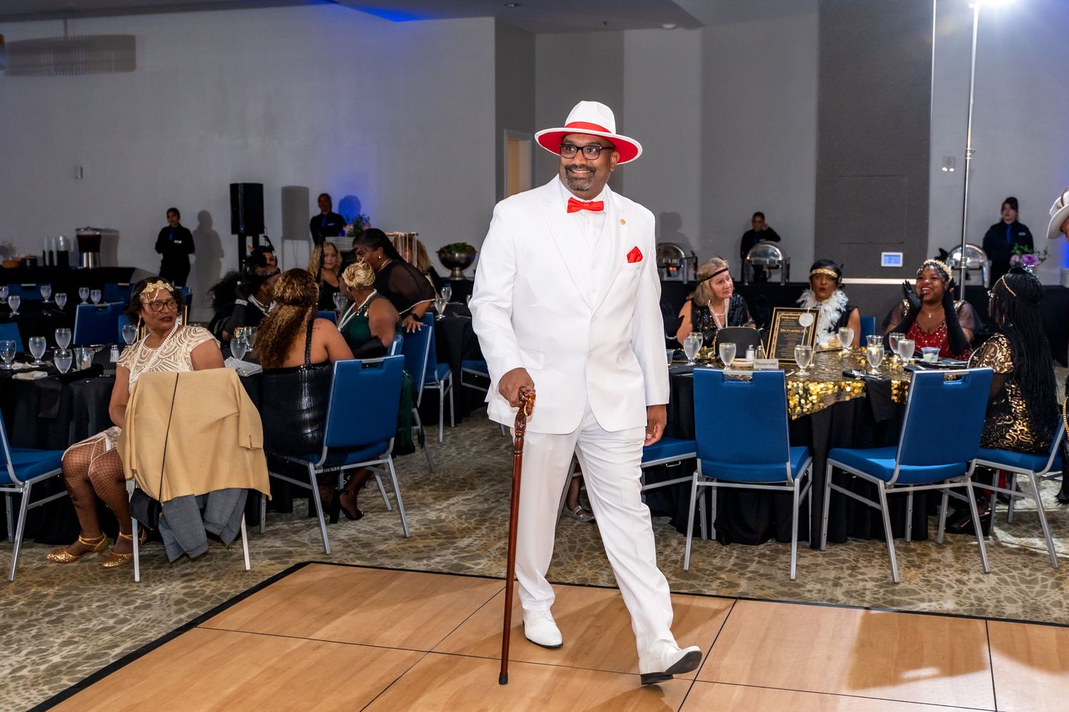 A person in an all-white suit and red bow tie dances on a wooden floor at a formal event.