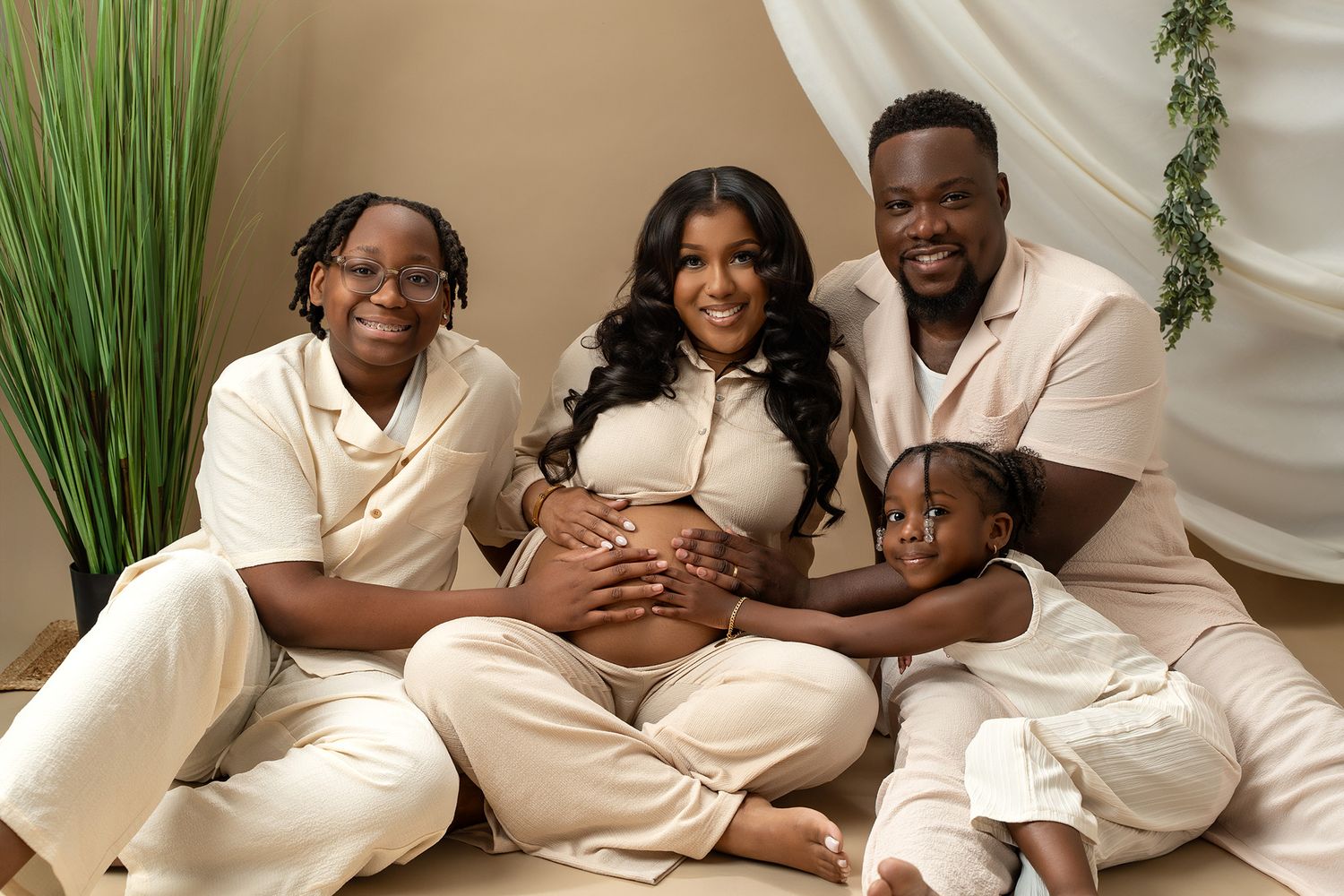 Family maternity photoshoot with parents and child wearing coordinating cream outfits against a beige backdrop.