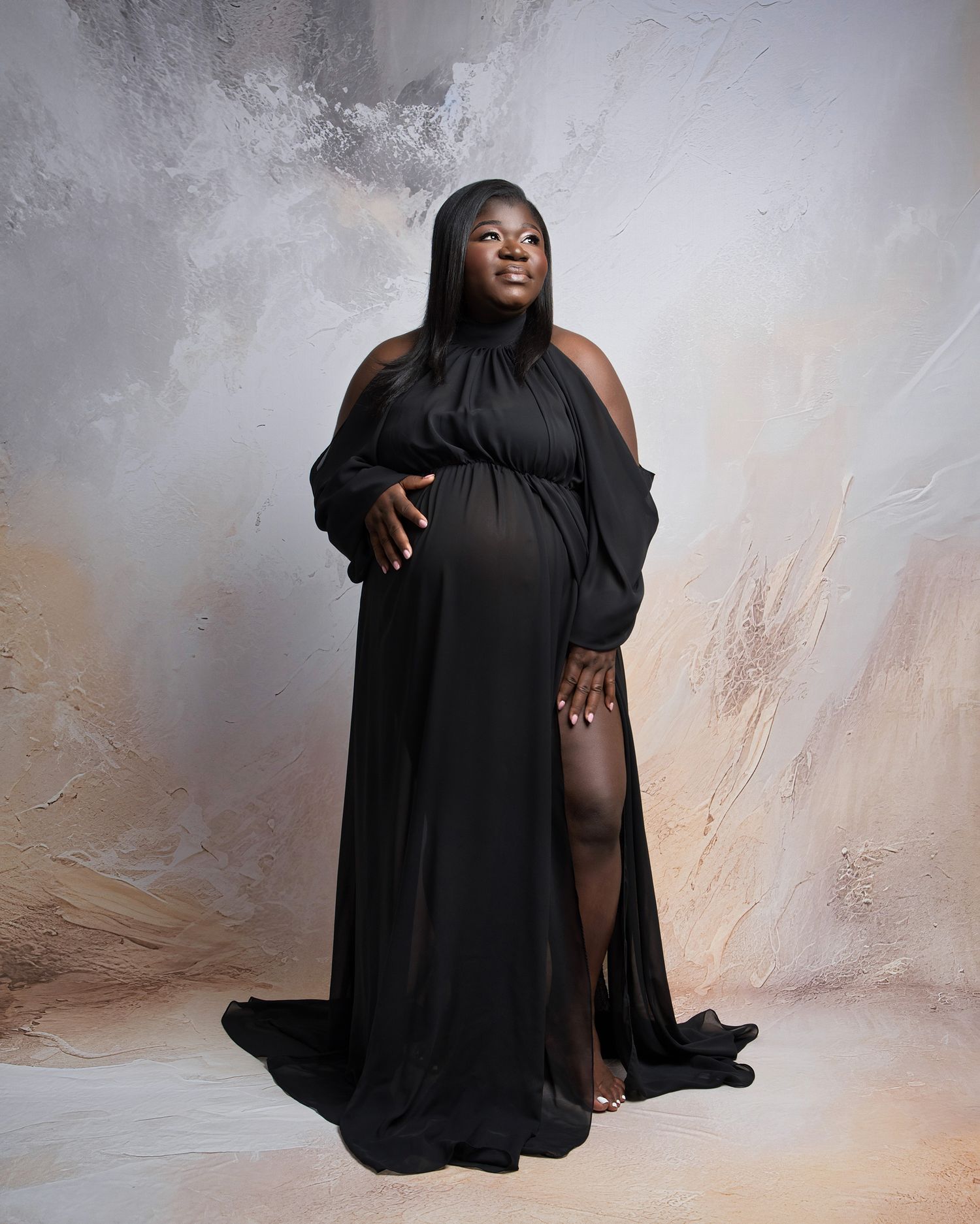 Dramatic maternity portrait in black flowing gown against textured neutral background.