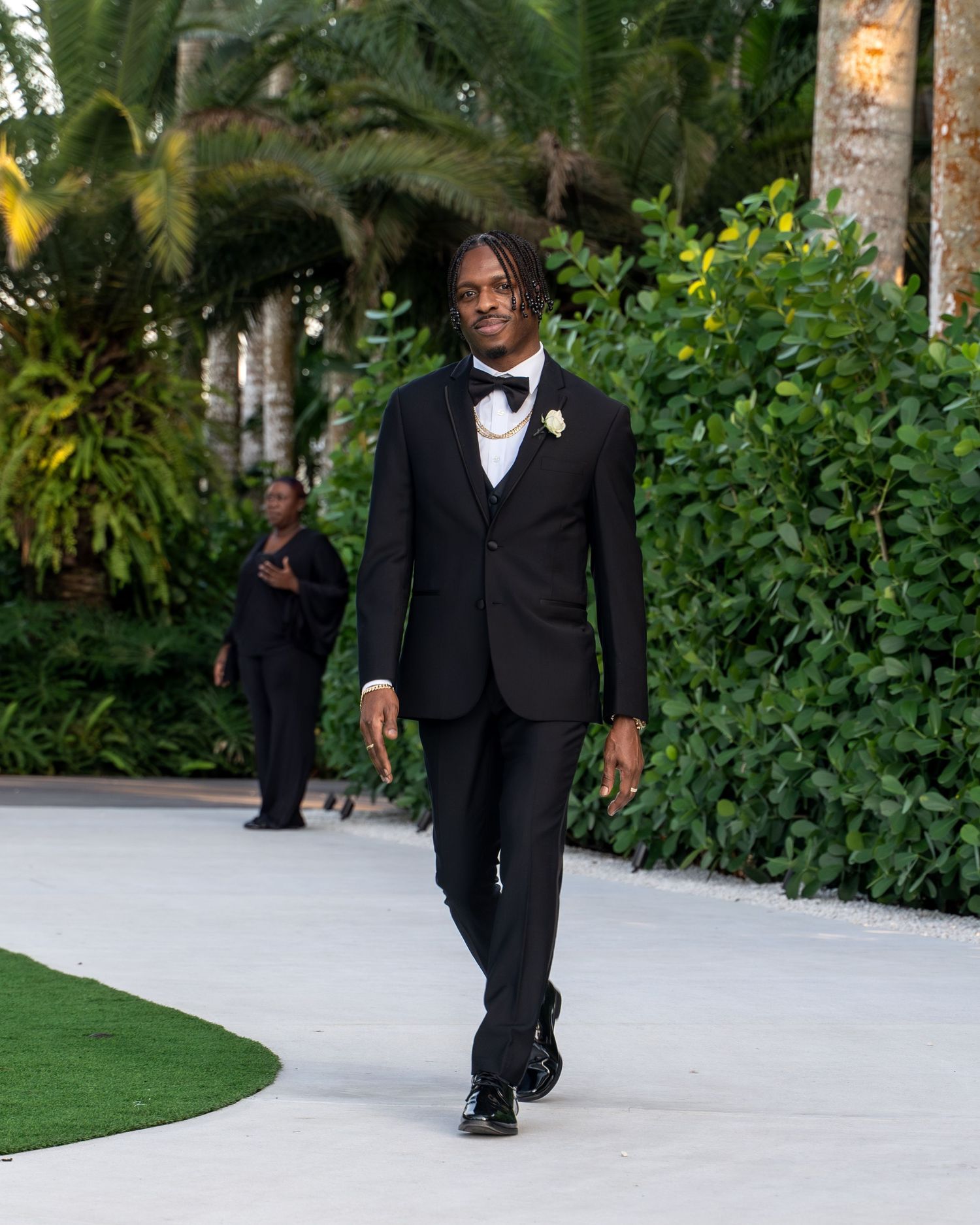 A person in a formal black tuxedo walks along a winding outdoor pathway surrounded by tropical landscaping.