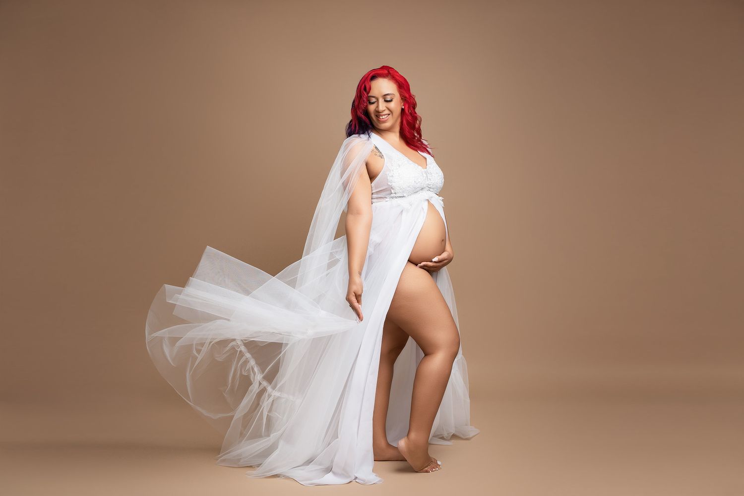 Maternity portrait in flowing white tulle dress against beige background.