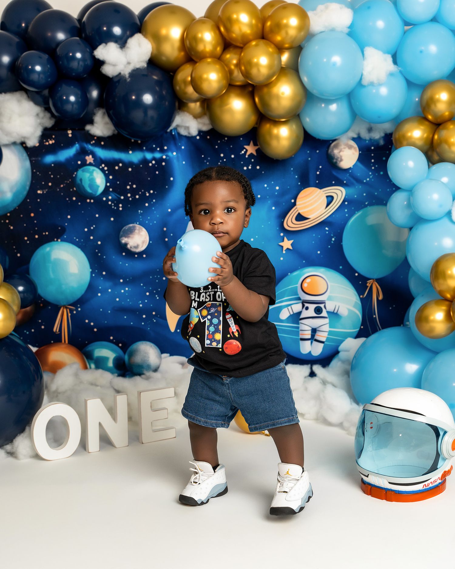 A space-themed first birthday photoshoot with navy, gold, and light blue balloon decorations and astronaut props.