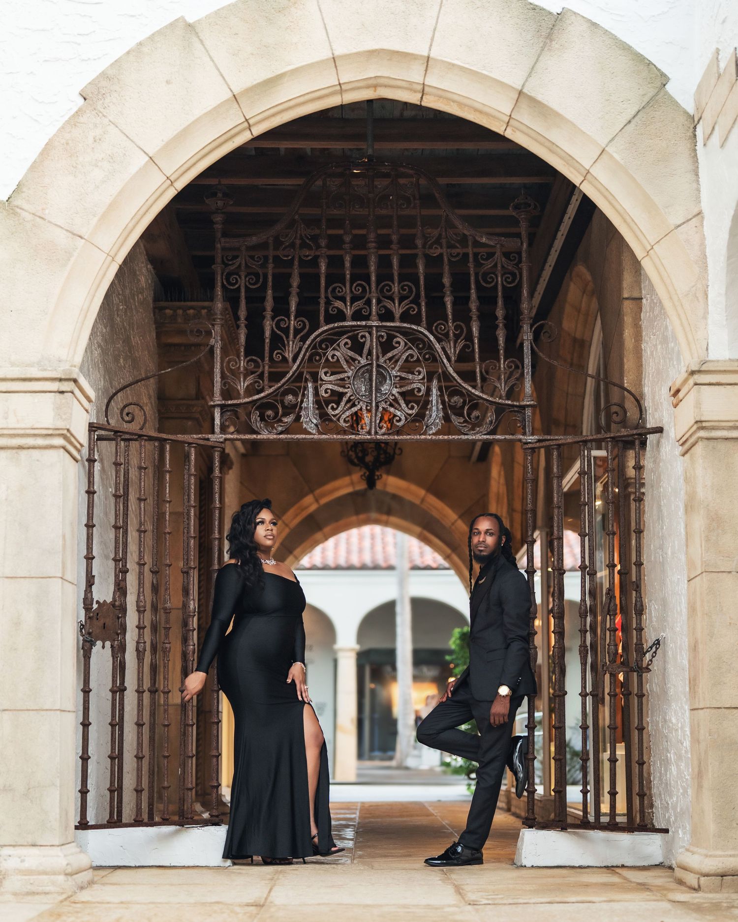 Two people in elegant black outfits pose beneath an ornate stone archway with wrought iron gates in a classical setting.