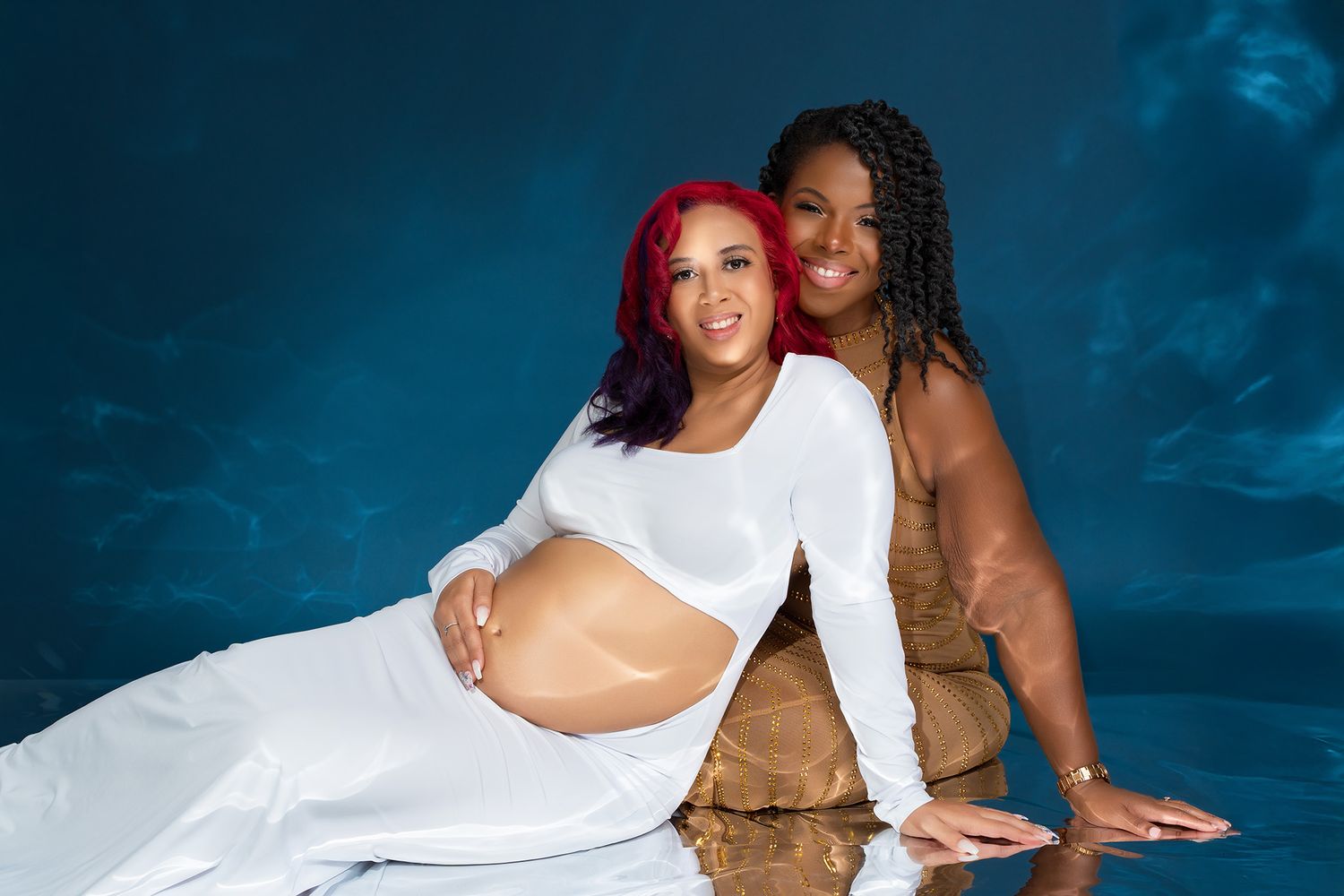 Two women pose together for a maternity photoshoot against a blue backdrop.