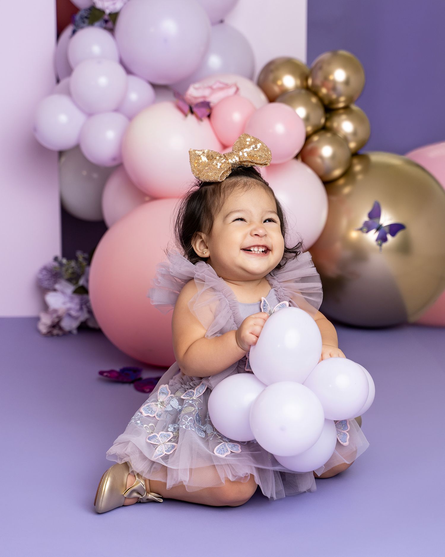 Festive first birthday photo scene with pink and gold balloons, butterflies, and white decorative elements.