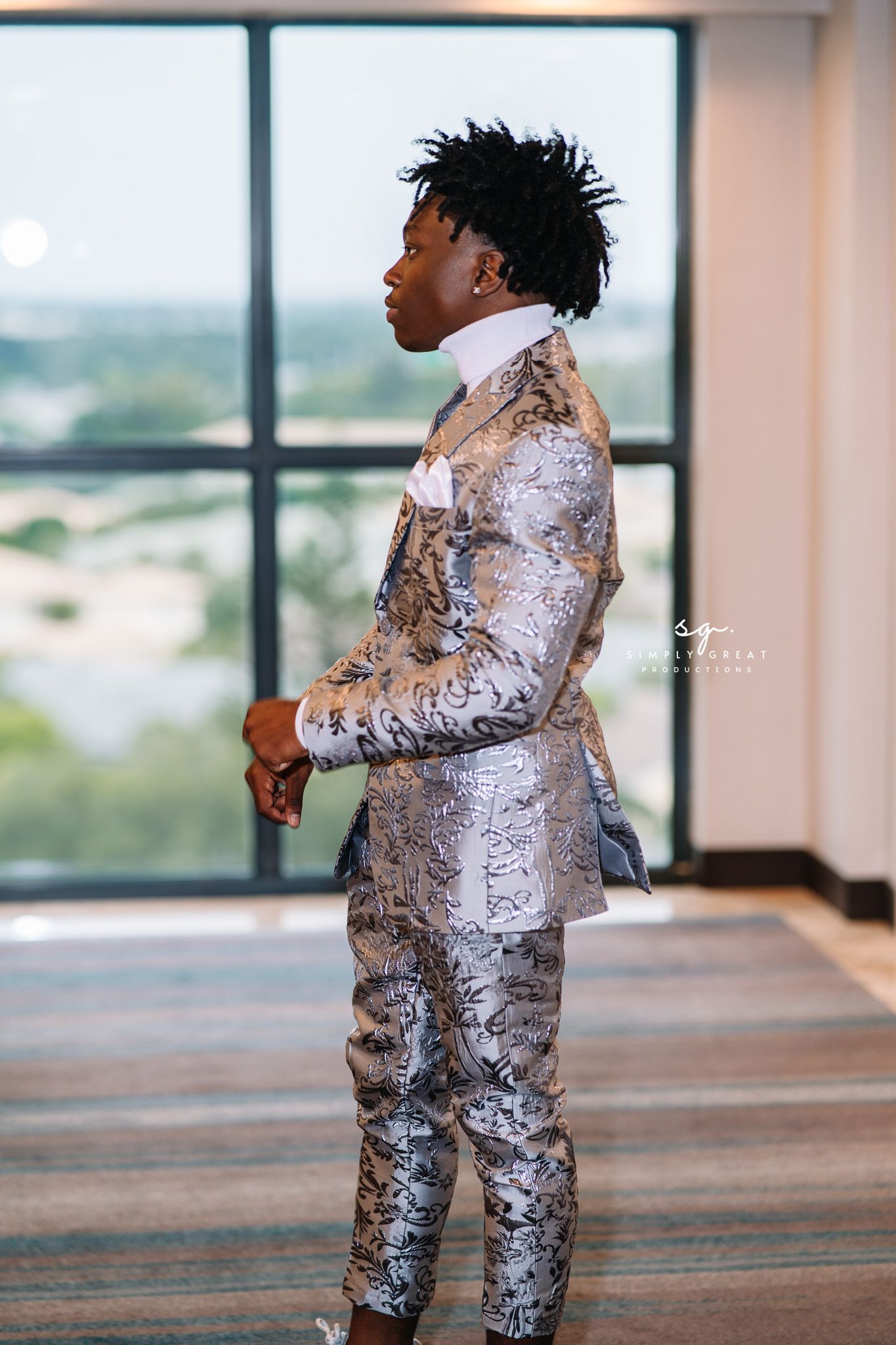 Person in silver patterned suit standing in profile view near large windows.