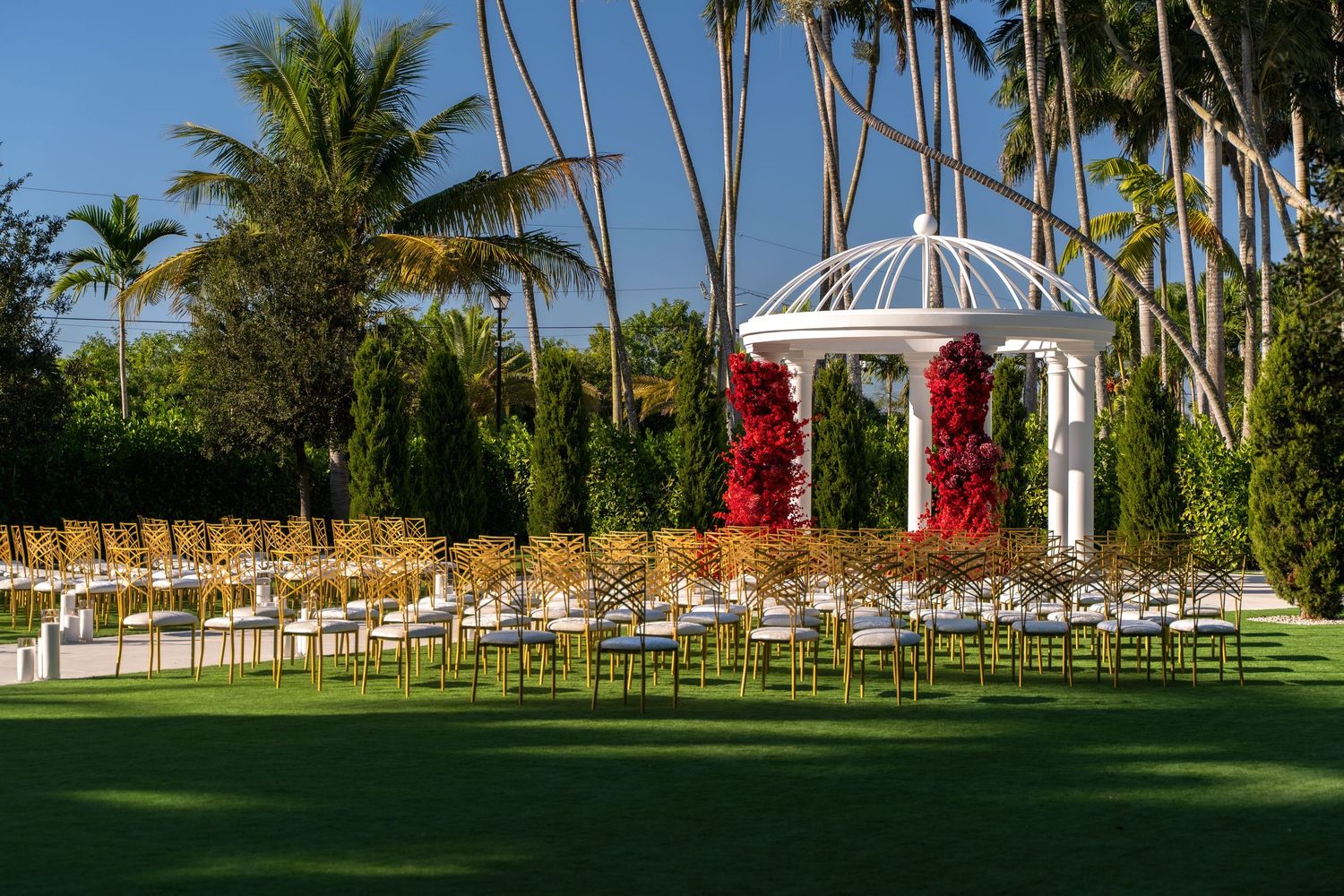 Panoramic view of a tropical wedding venue with palm trees, gazebo, and wooden chairs set up on manicured green lawn.