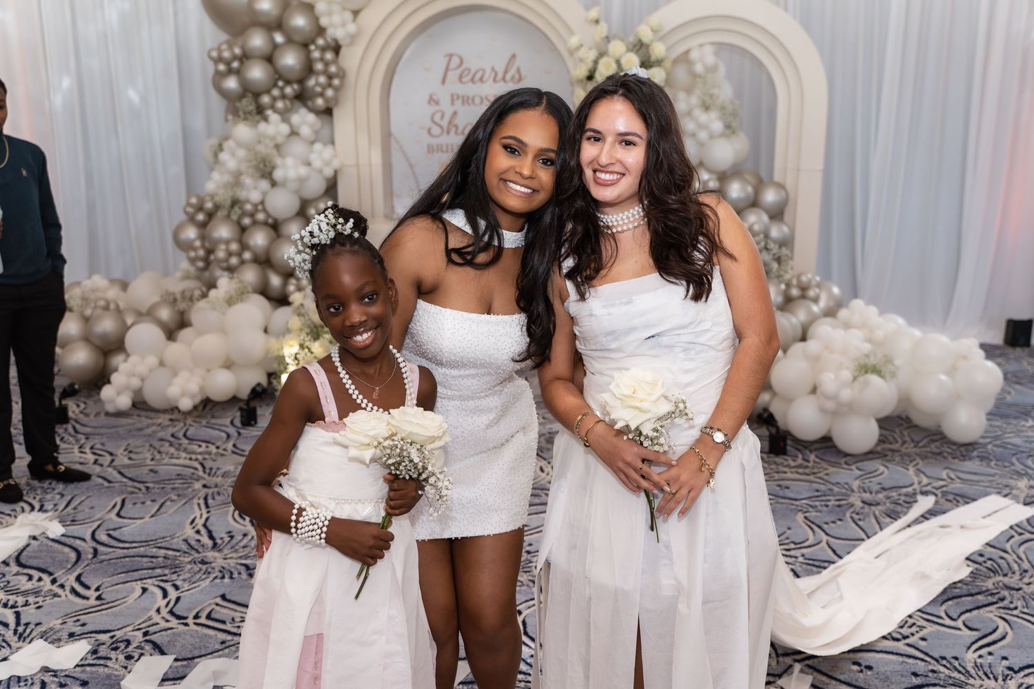 A diverse group of friends pose together at a glamorous white and silver themed party celebration with balloon decor.