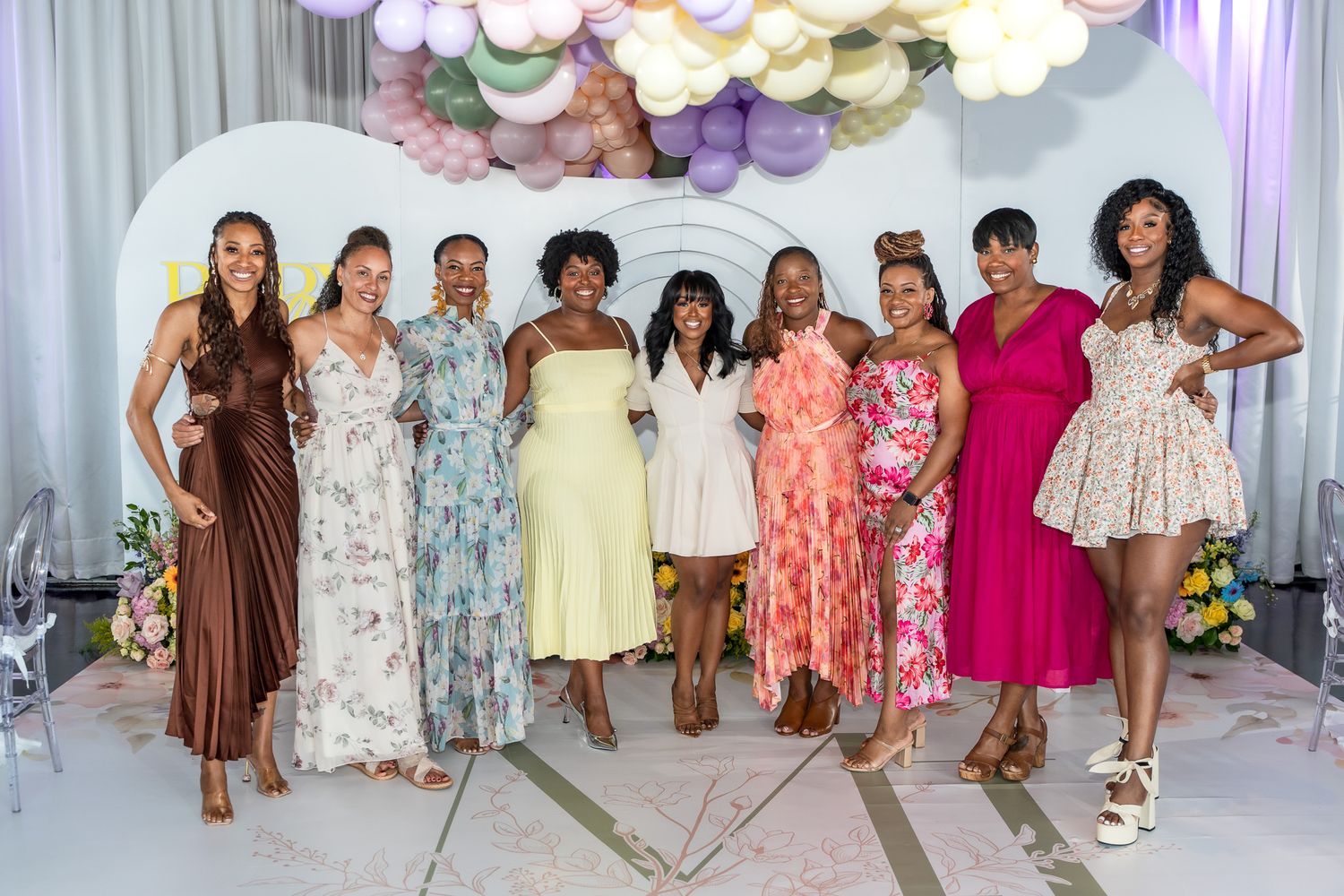 A group in colorful dresses poses together under pastel balloon decorations at an elegant party venue.