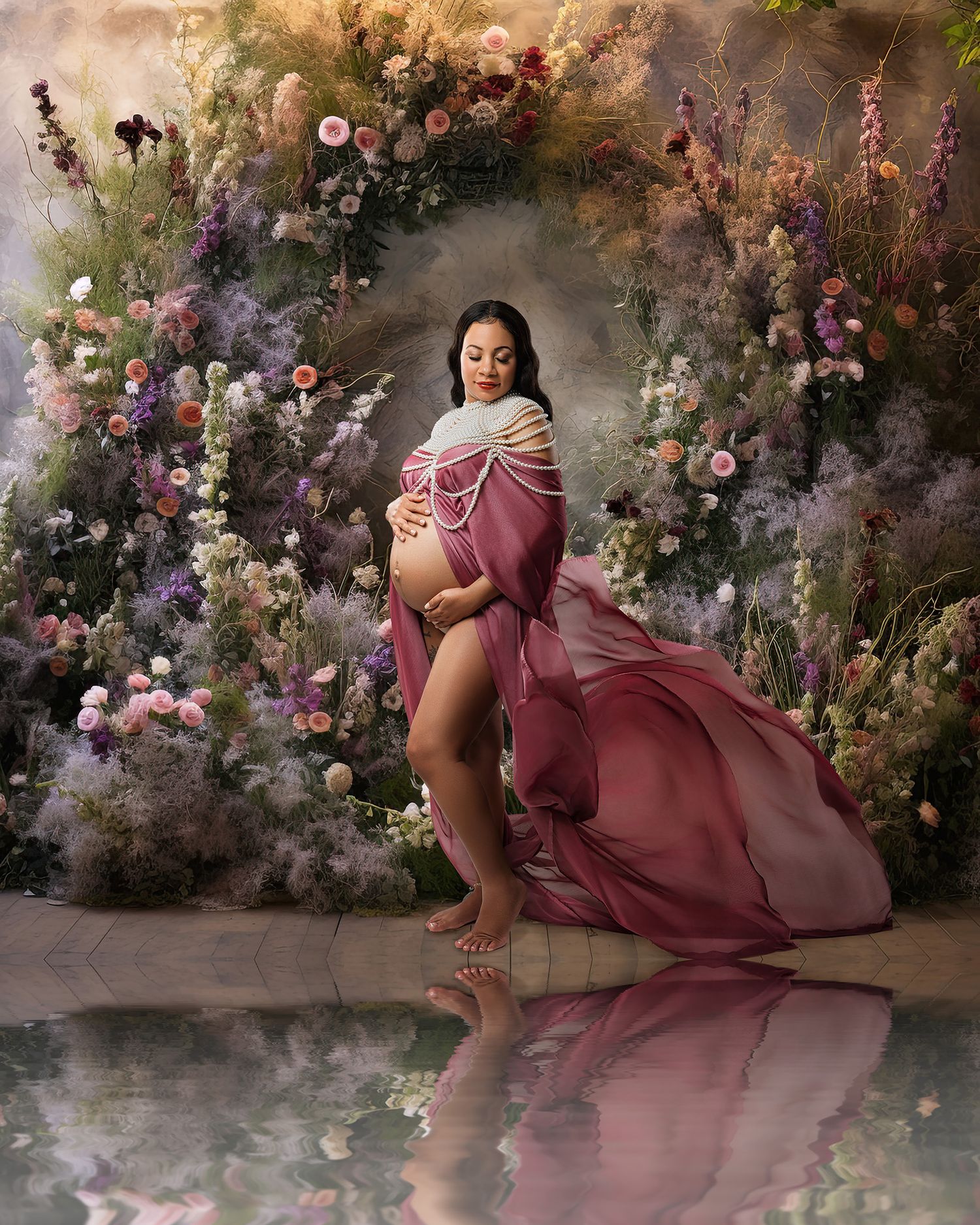 Romantic maternity photo with floral arch backdrop and flowing pink dress creating dreamy atmosphere.