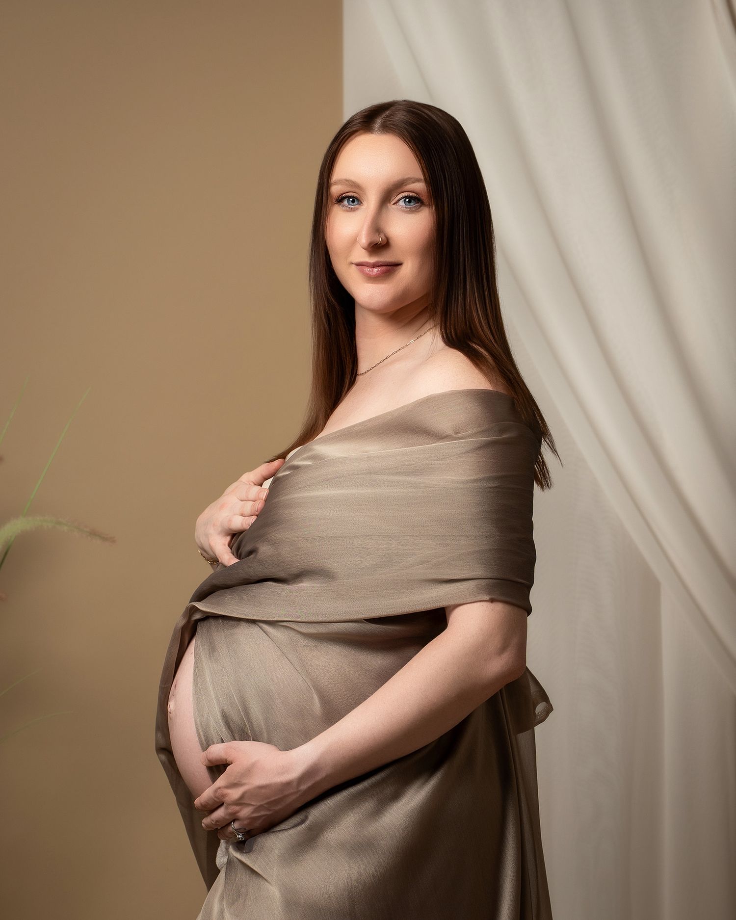A maternity photo featuring a woman in a draped taupe fabric against a neutral backdrop with soft lighting.