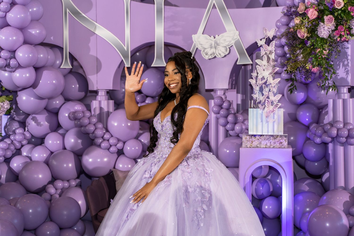 A celebrant in a lavish purple and white gown poses against a balloon arch backdrop.