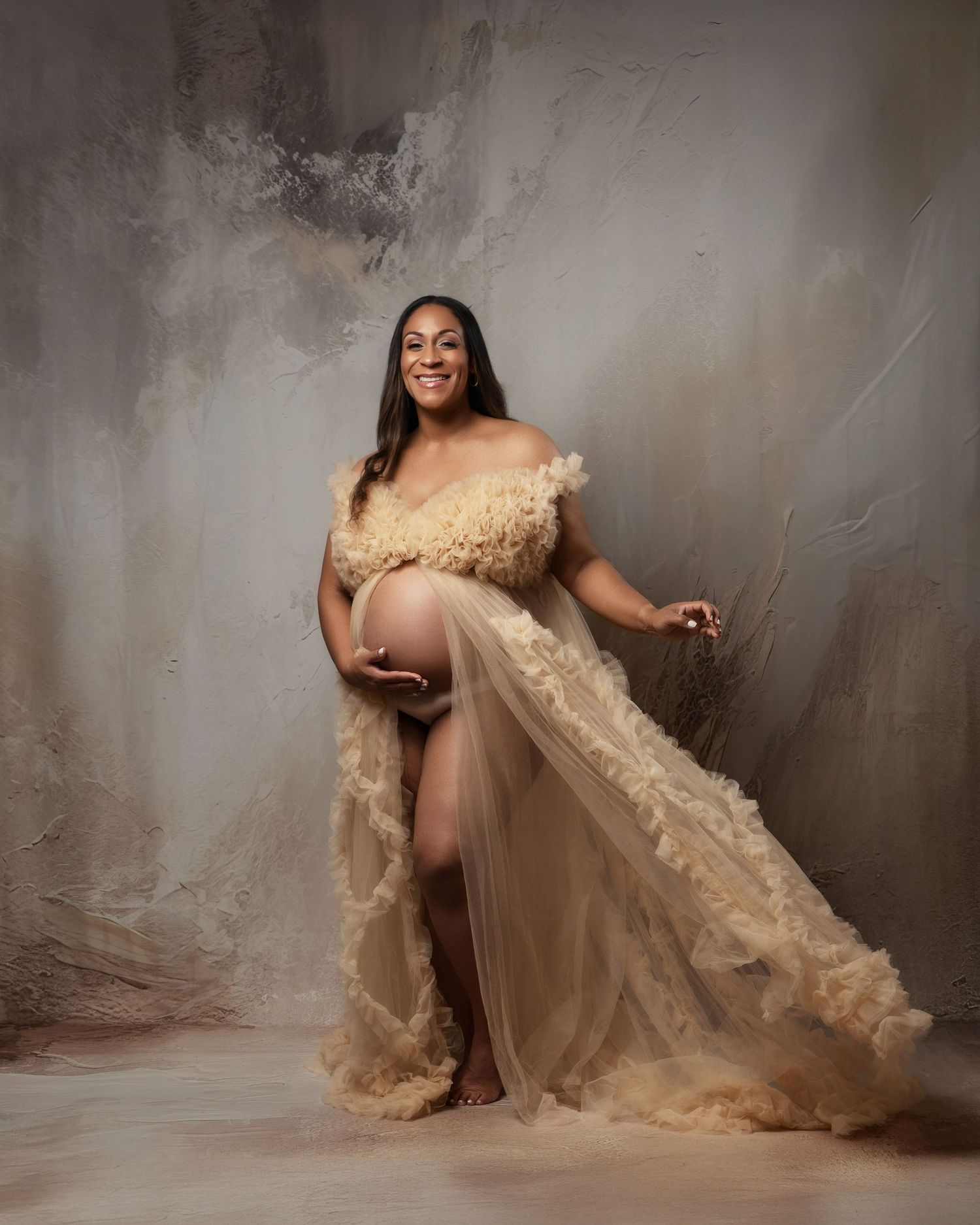 A glamorous maternity photoshoot featuring a flowing beige gown against a textured grey backdrop.