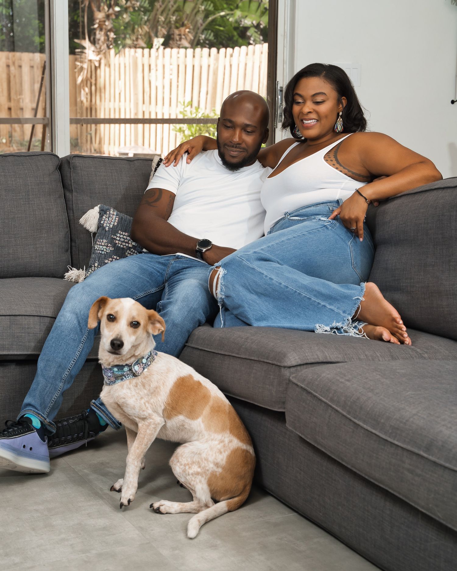 A couple relaxes on their gray sofa with their tan and white dog.