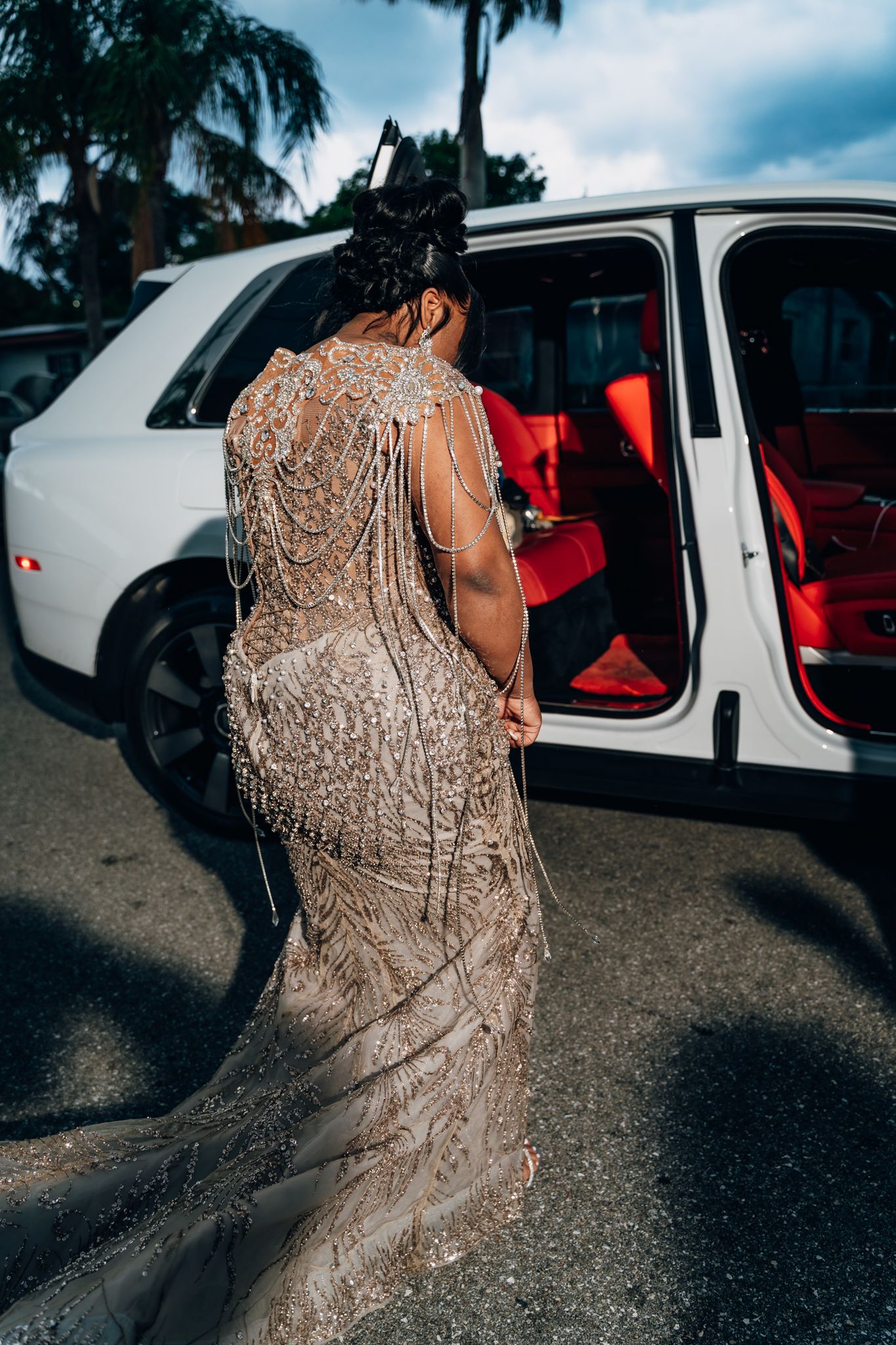A stunning beaded gown with intricate back details as someone enters a white luxury vehicle with red interior.