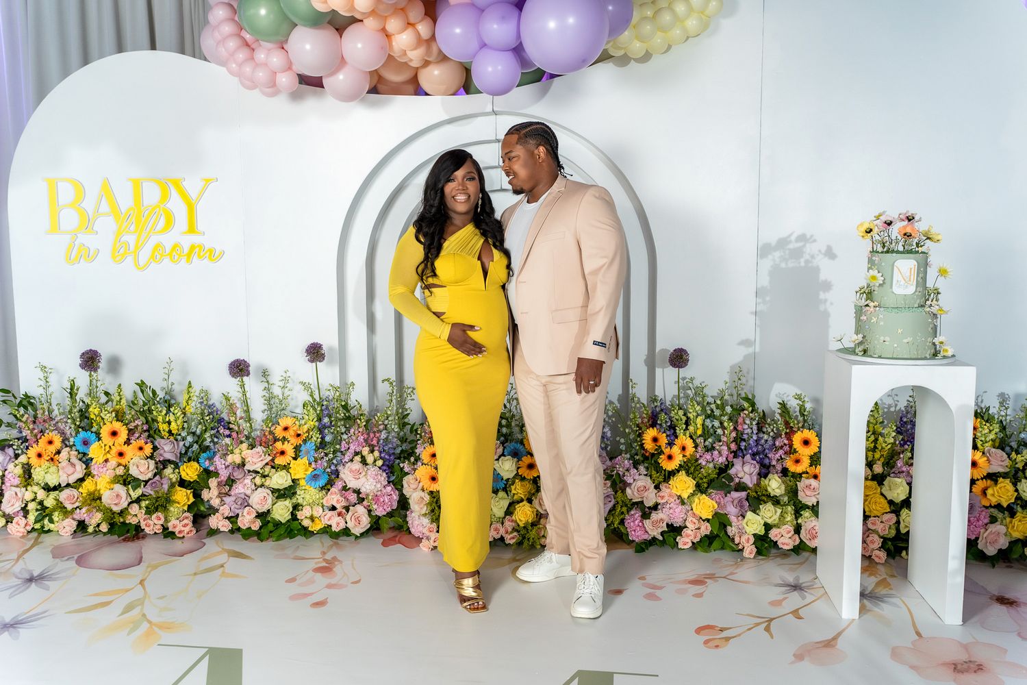 Expectant couple standing in coordinated outfits against floral arch and balloon display.