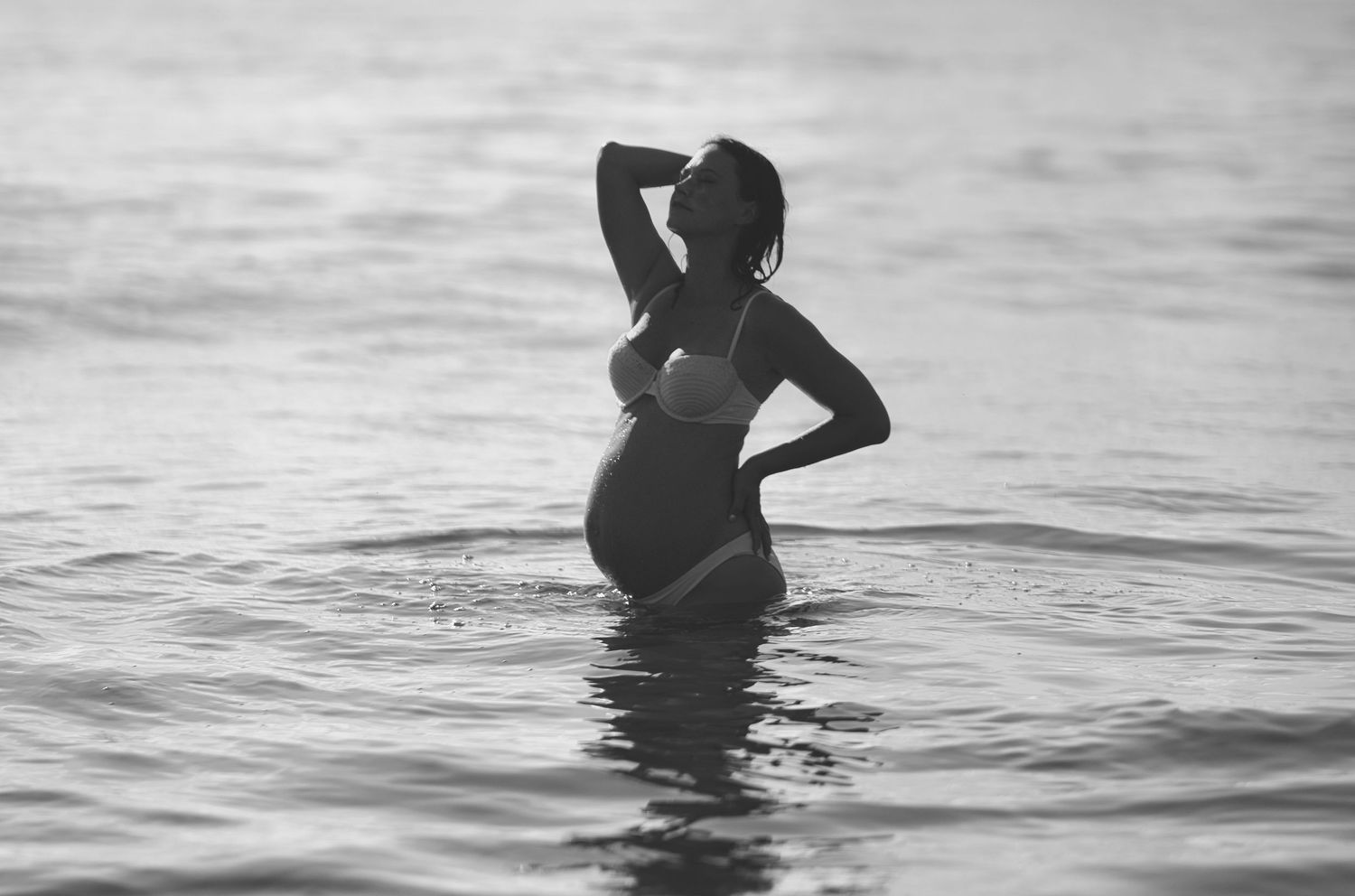 Silhouette of pregnant woman standing in calm ocean water during sunset.