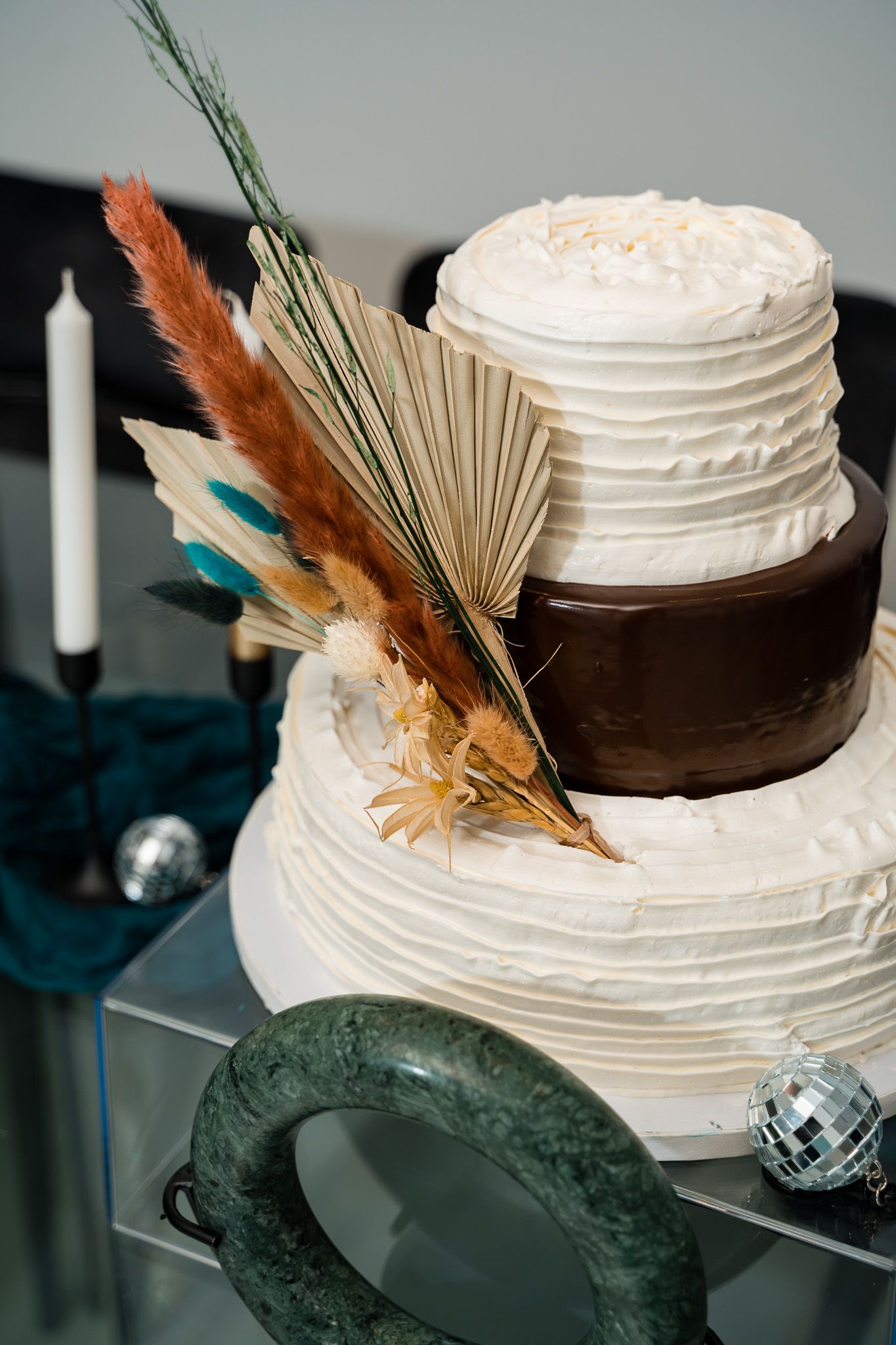 Two-tiered wedding cake decorated with dried palm leaves and feathers in white and chocolate colors.