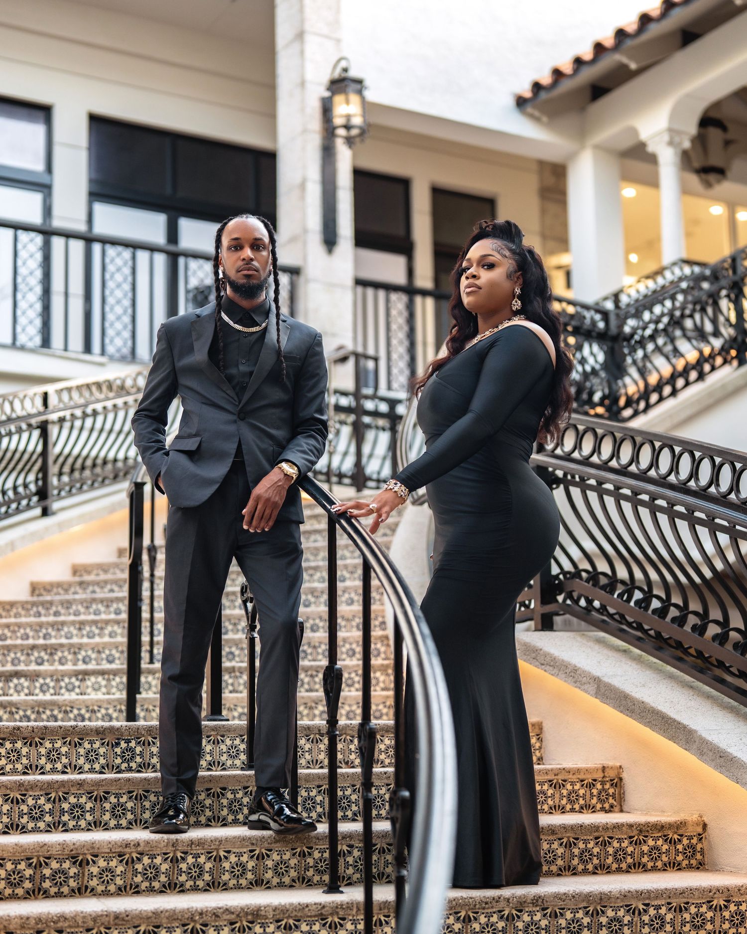An elegant couple poses on an ornate Mediterranean-style staircase with decorative wrought iron railings.