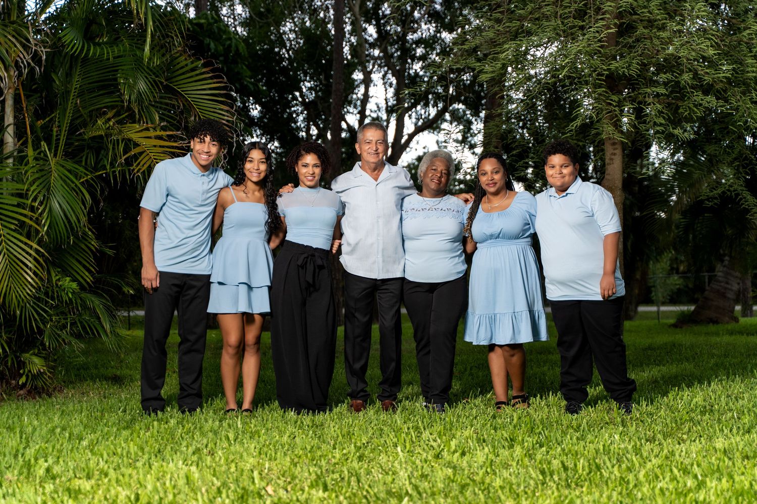 A group in coordinating blue and white outfits poses together on a grassy lawn with palm trees in the background.
