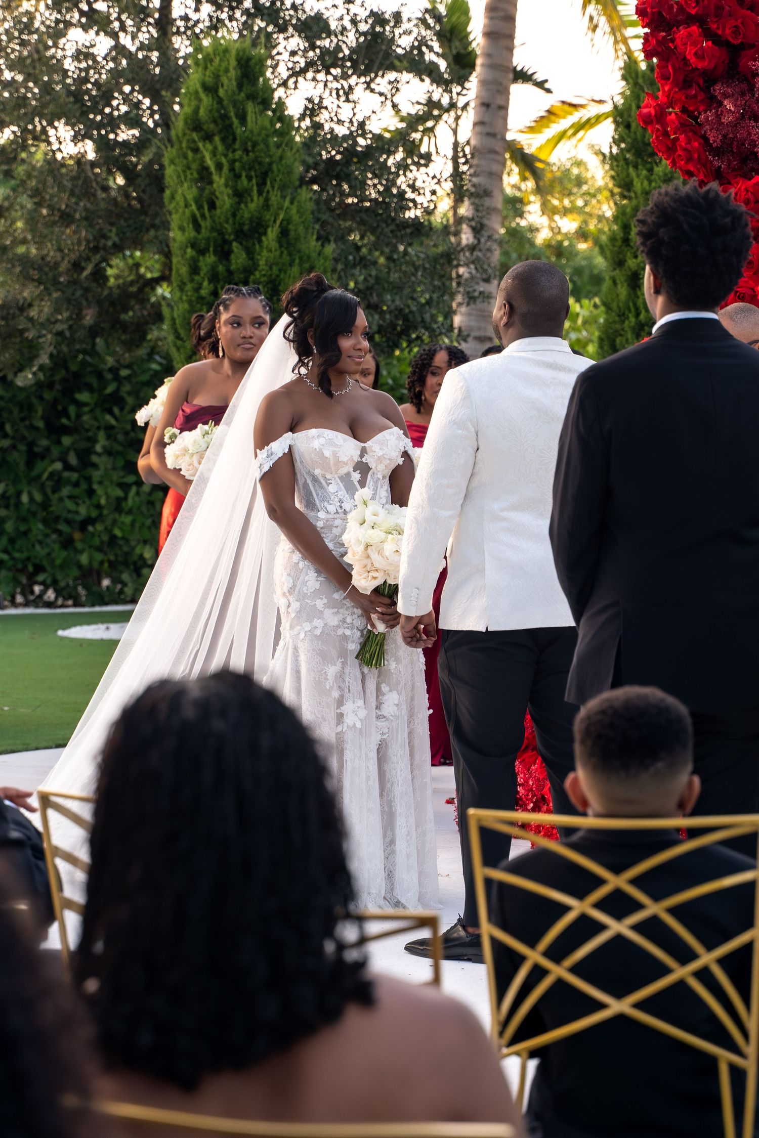 An outdoor wedding ceremony with guests seated on gold chairs while the couple exchanges vows under palm trees.