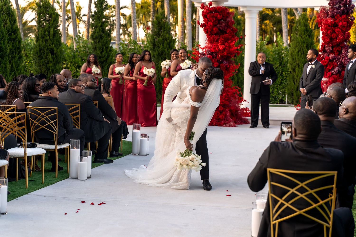 A wedding ceremony with red floral decor, gold chairs, and guests watching the couple's first kiss.