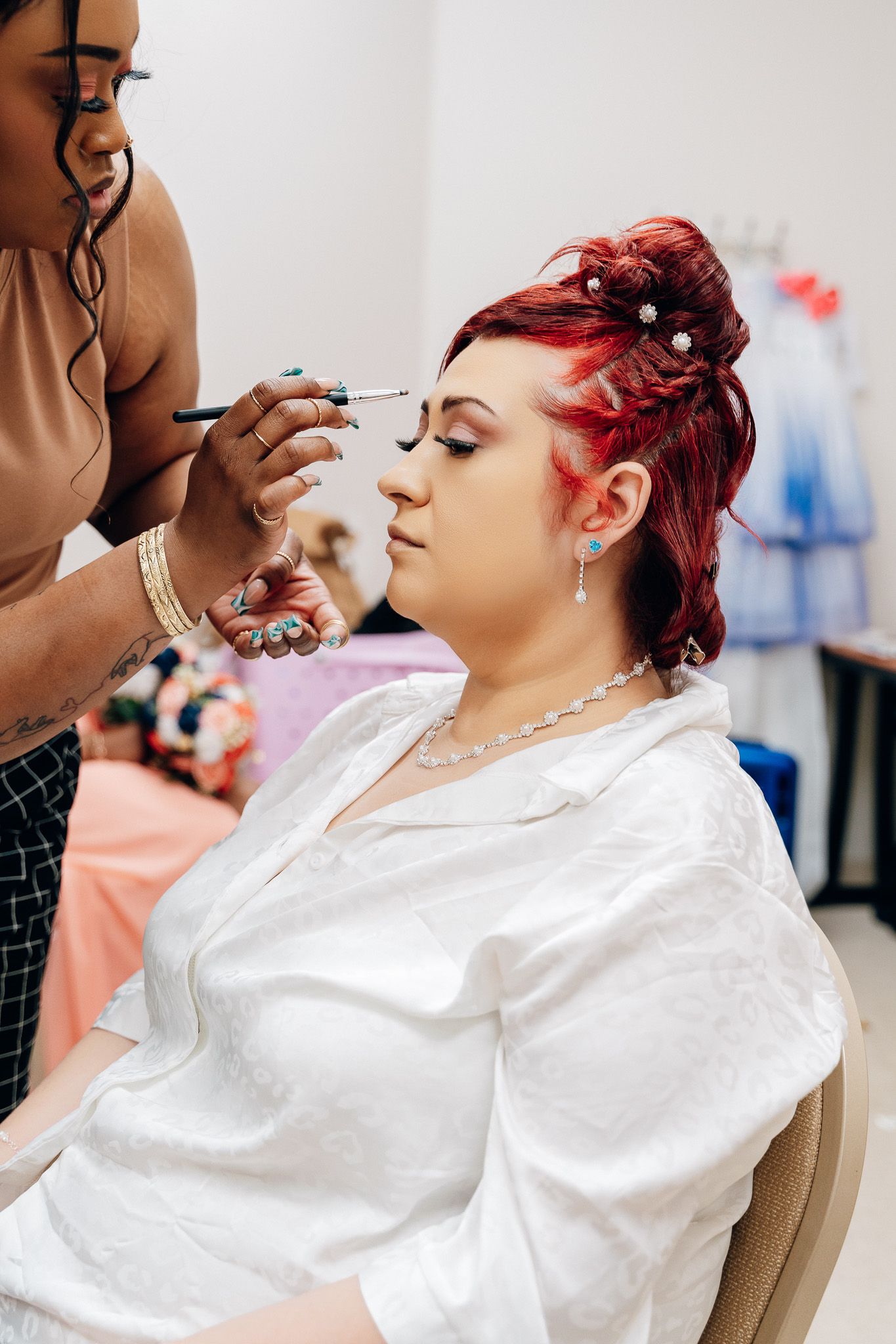 A makeup artist applies eye makeup to a bride with vibrant red hair.