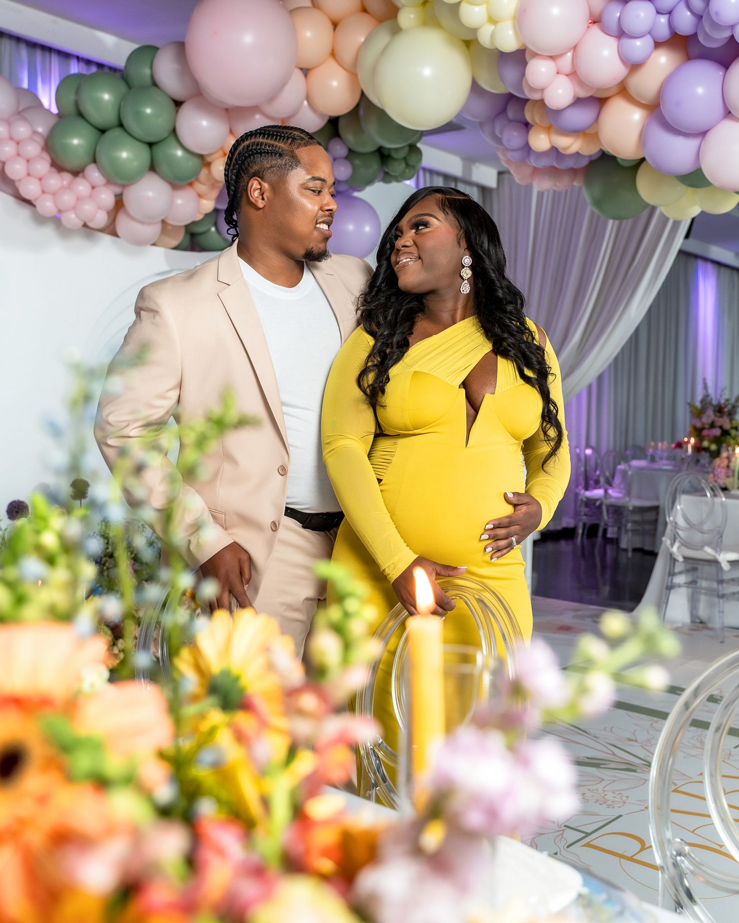 Maternity photo shoot with couple in yellow dress and beige suit amidst floral decorations and balloons.