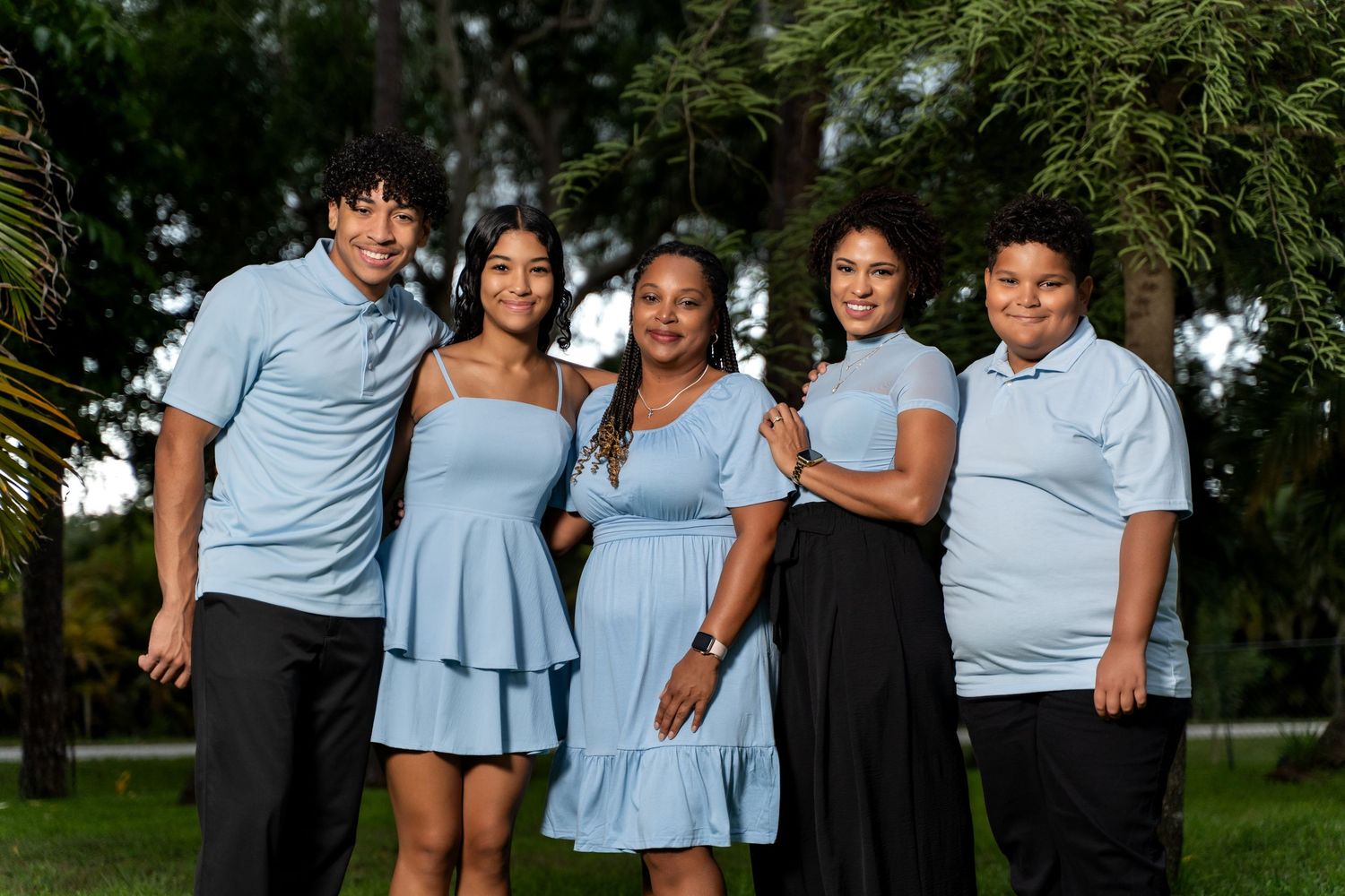 A family wearing coordinated light blue outfits poses together for outdoor portrait photos in a park setting.