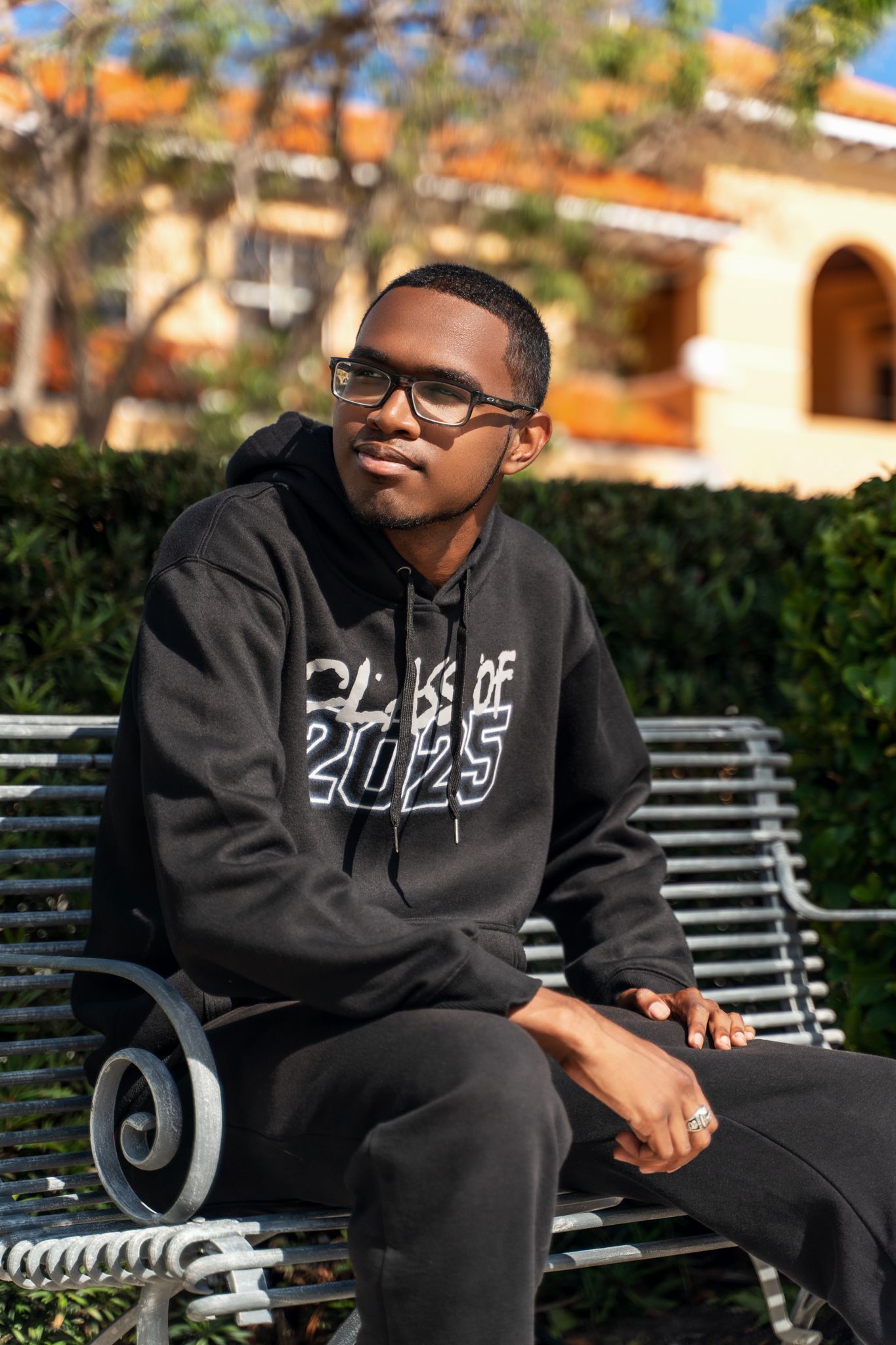 A person in glasses wearing a black Class of 2025 hoodie sits relaxed on an ornate metal bench in a garden setting.