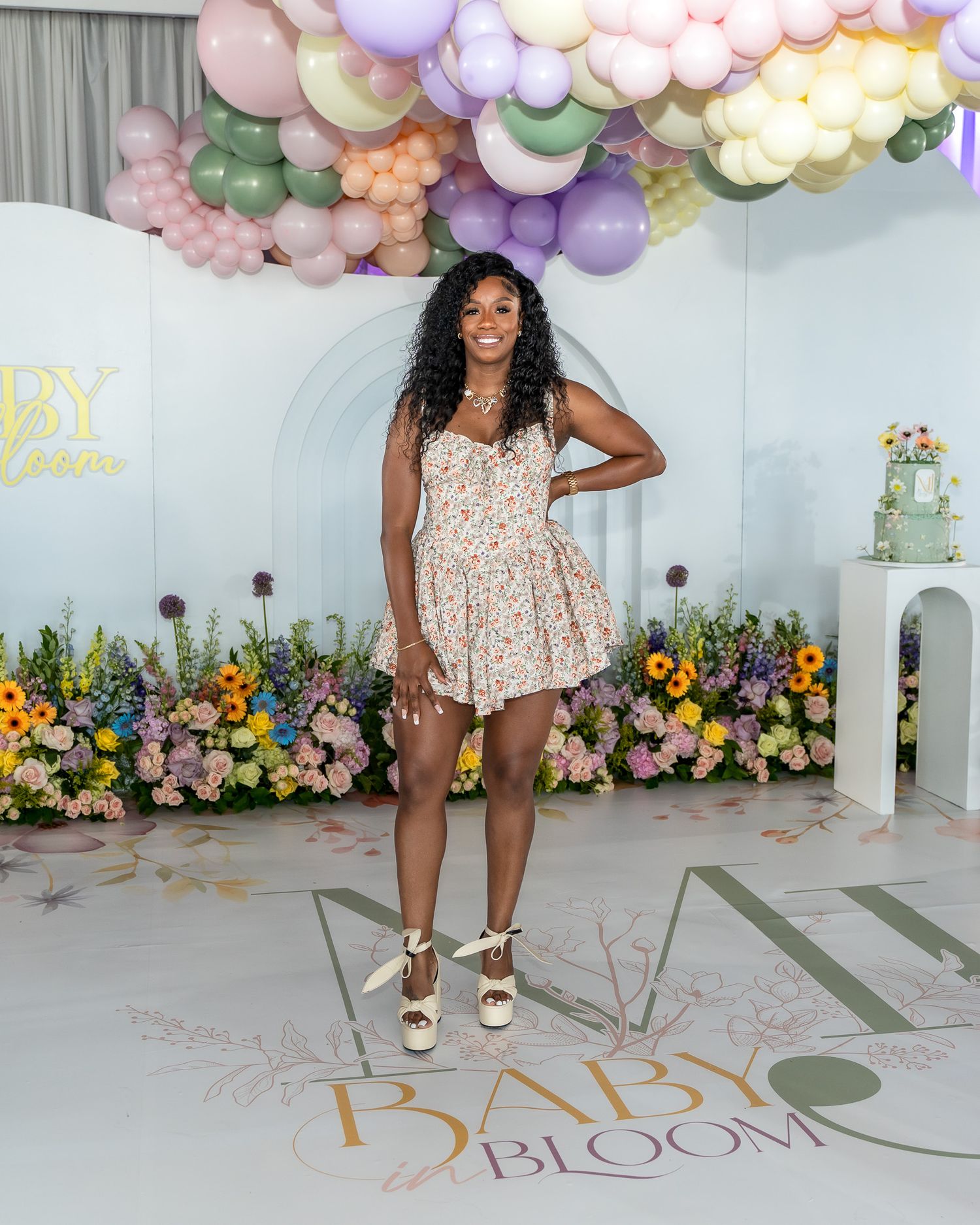 A guest poses under a pastel balloon arch with floral decorations at a special event.