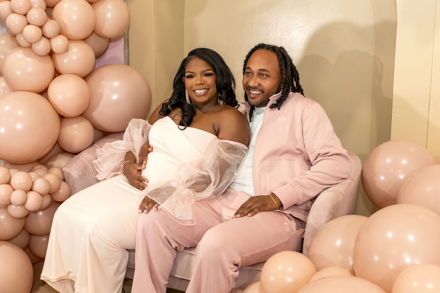 Couple in coordinating pink and white outfits pose together against balloon backdrop at celebration.