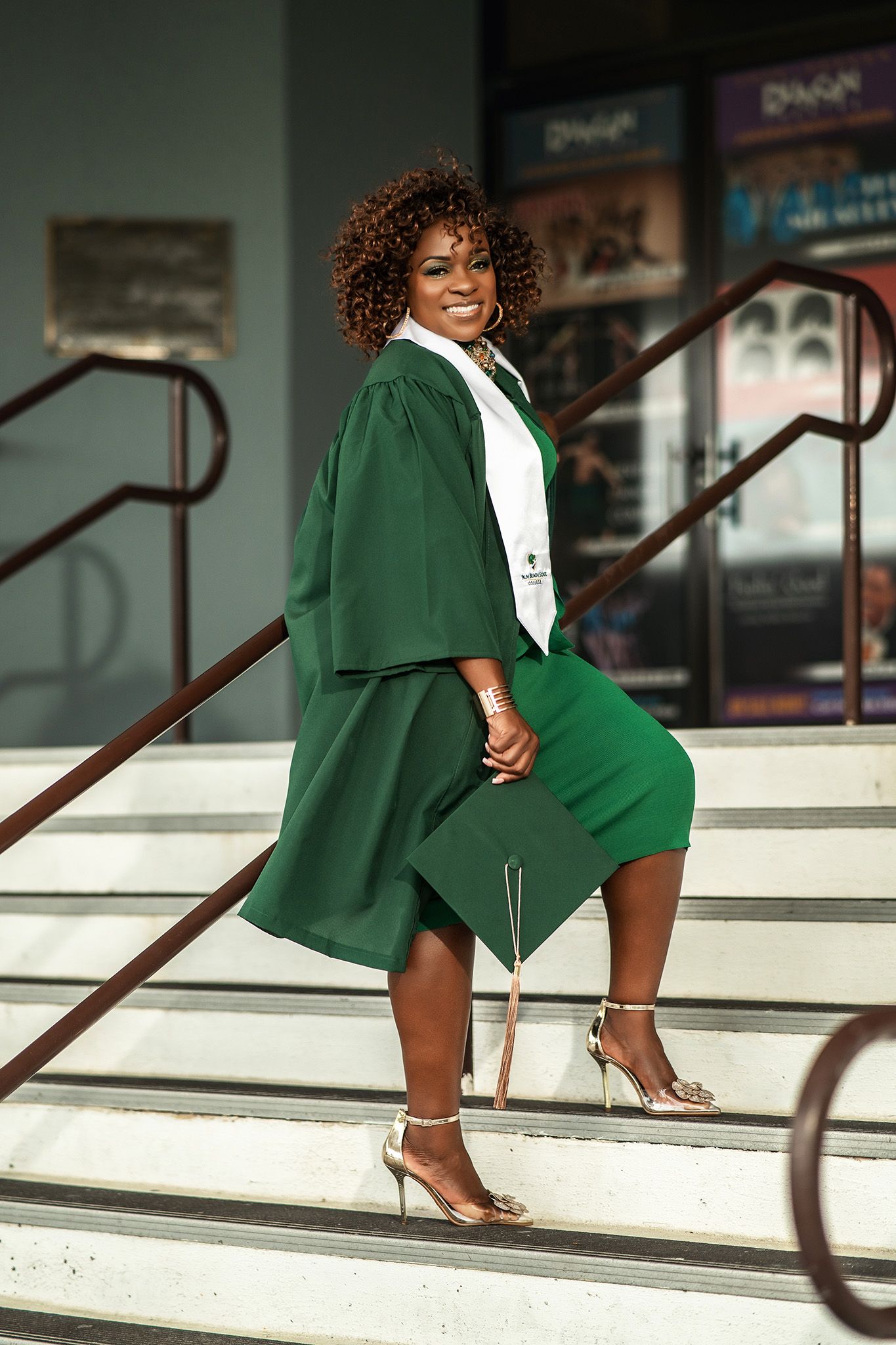 Graduate in green academic regalia poses joyfully on stairs with railing.
