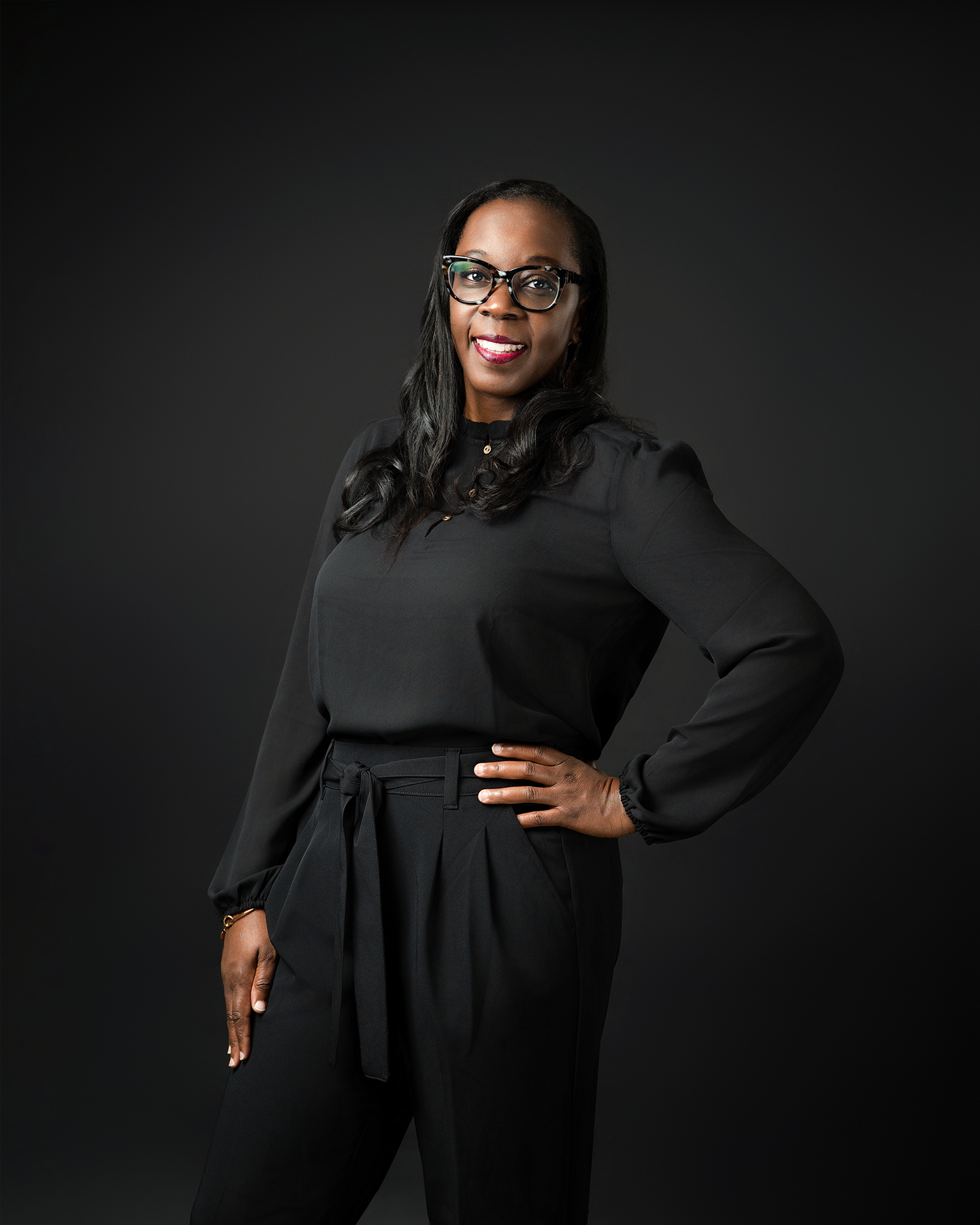 A professional portrait in black attire against dark background with dramatic lighting.